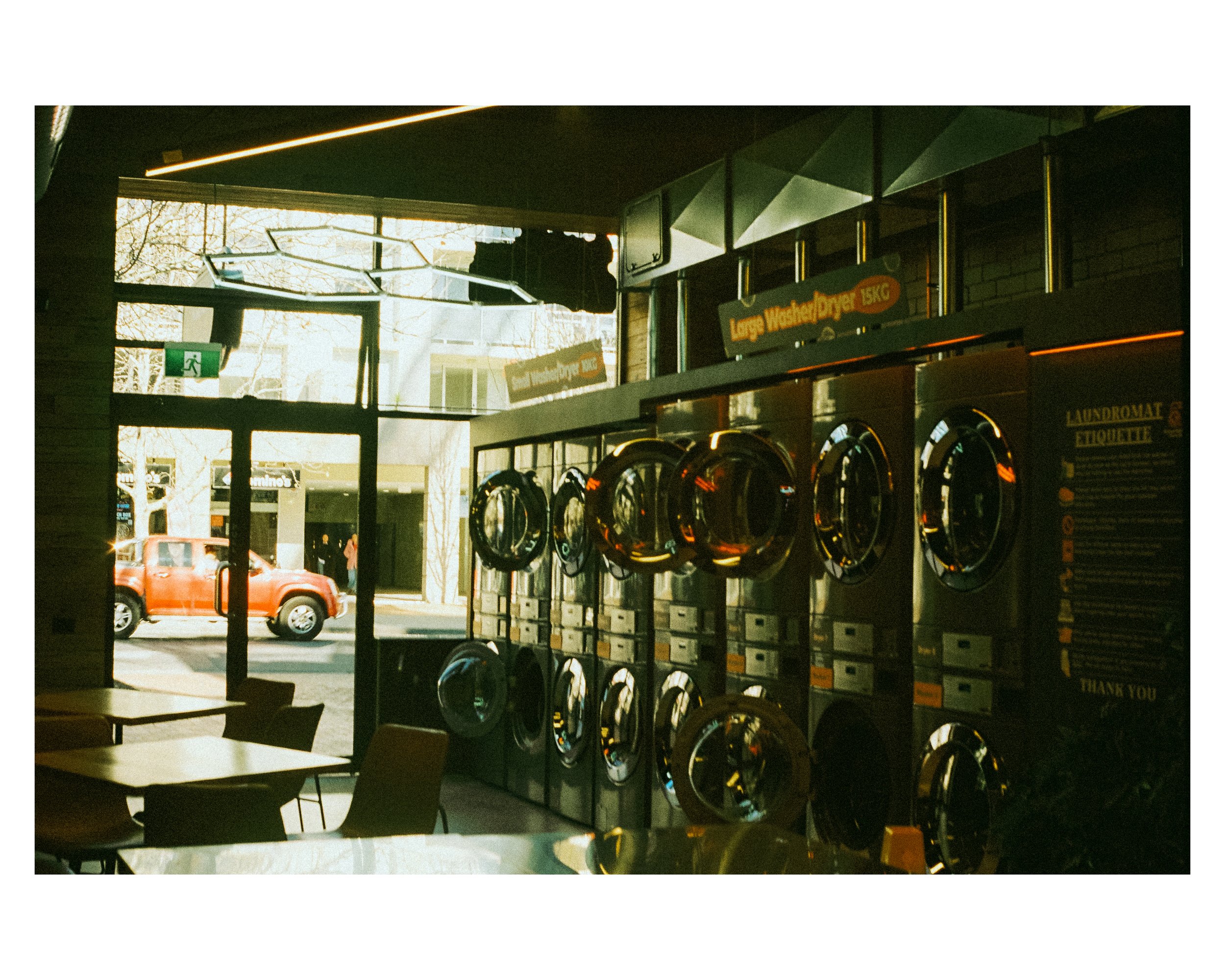 Interior view of a laundromat with washing machines, tables and chairs, and large windows showing a street scene outside with a red pickup truck