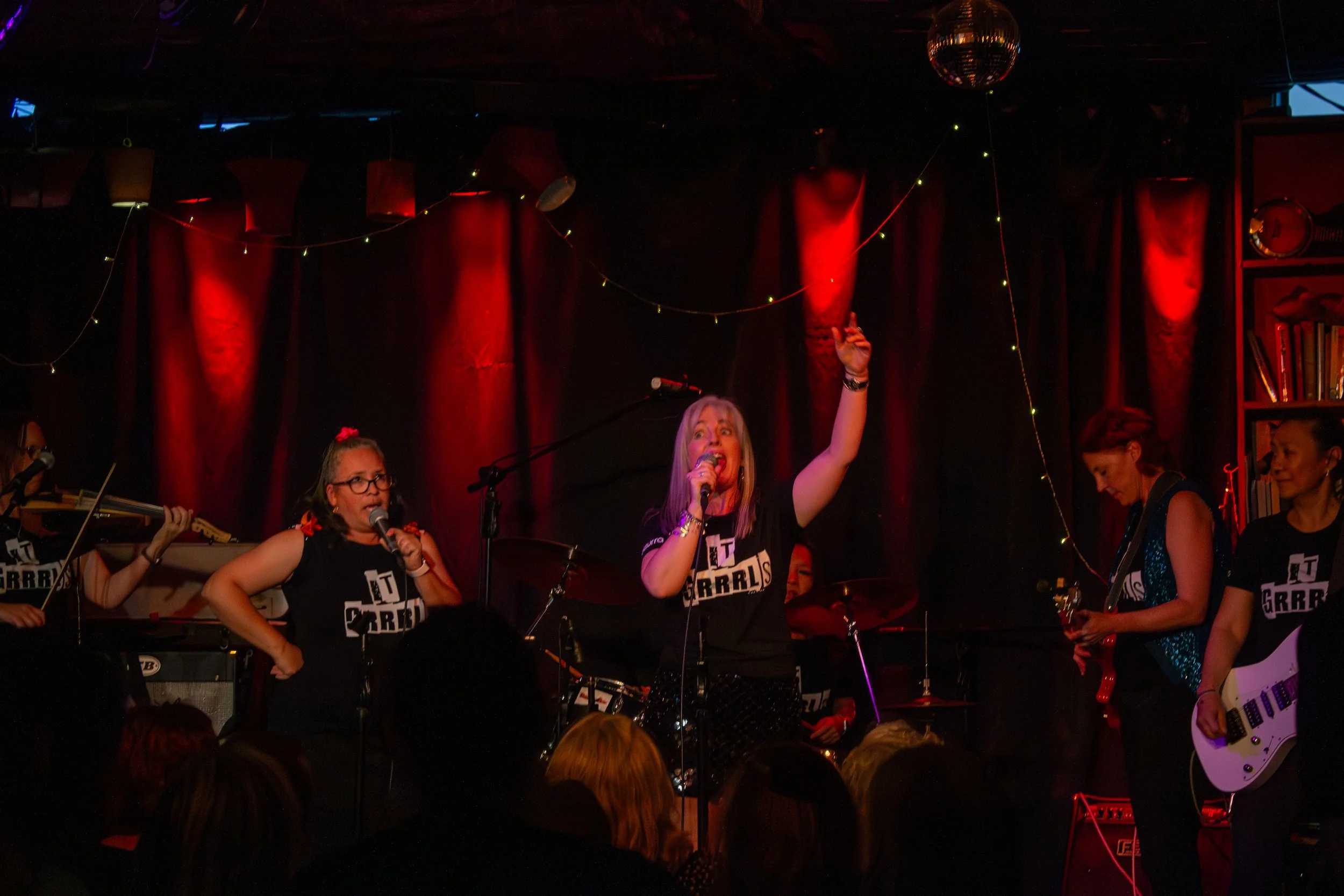 Women performing on stage with musical instruments and microphones, red stage curtains, string lights, and a disco ball overhead.