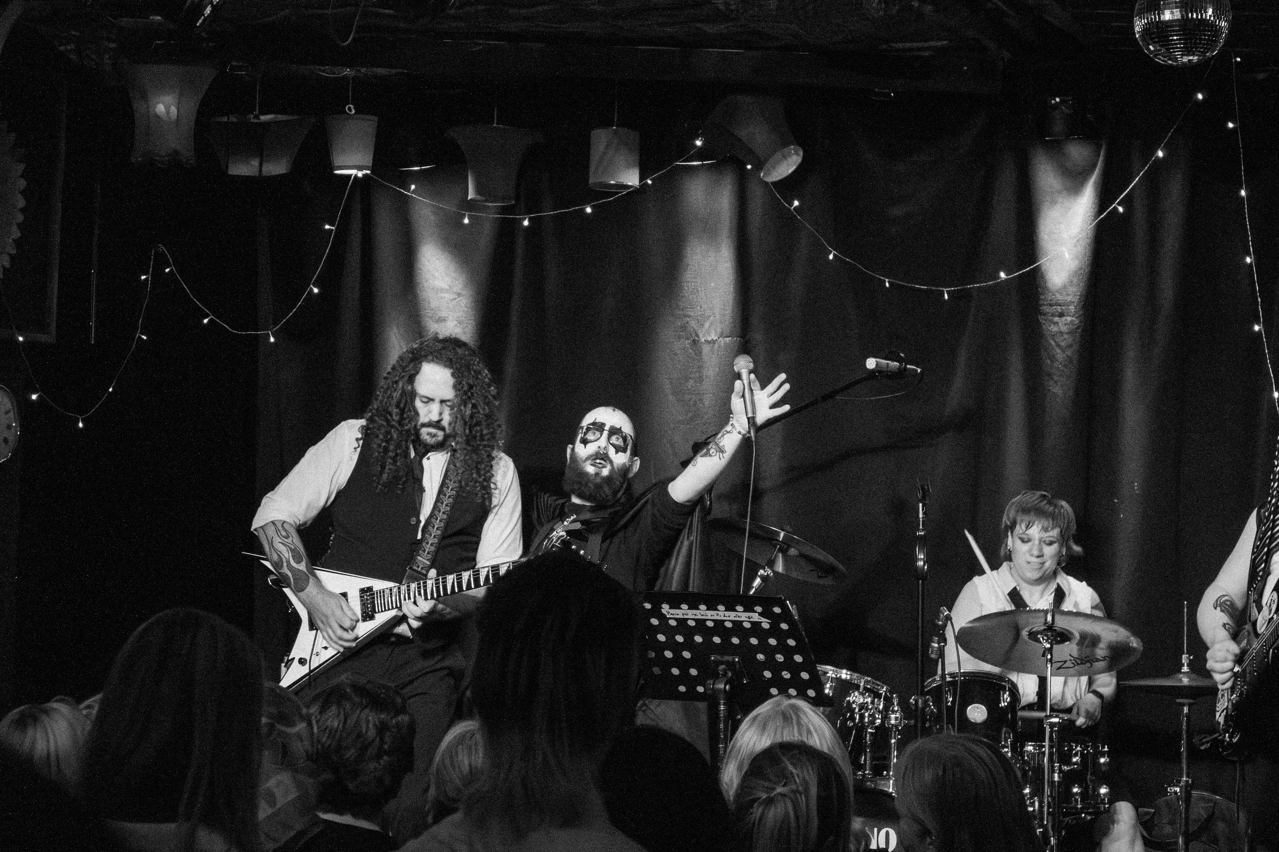 A band performs live on stage, featuring a guitarist, a drummer, and another guitarist, with an audience in front, and stage decorations of hanging lights and black curtains.