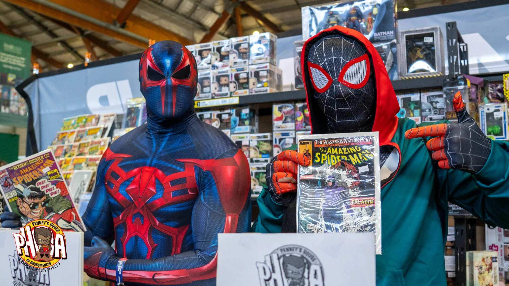 Two people dressed in Spider-Man costumes at a comic convention booth, with comic books displayed in the background