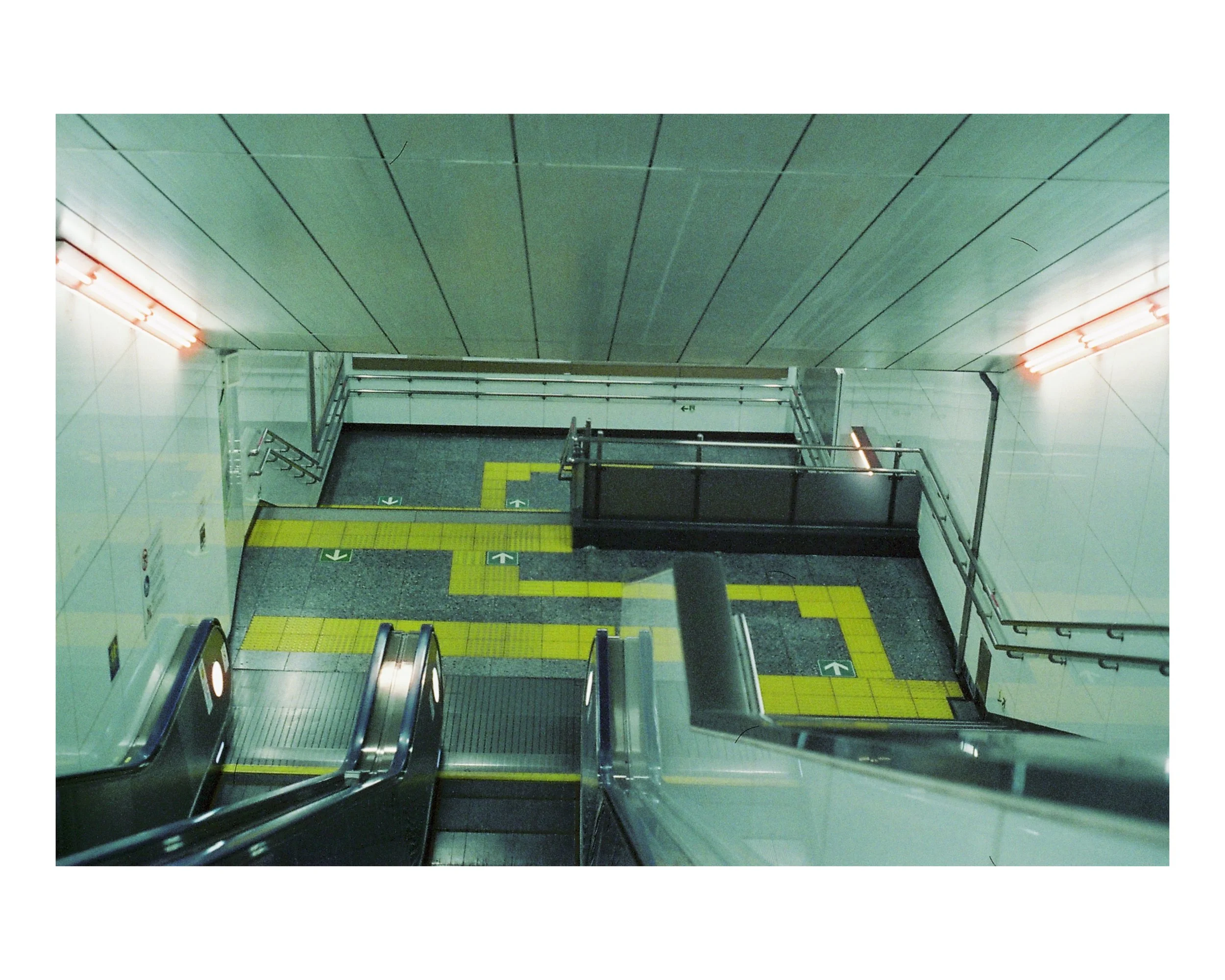 View from the top of an escalator in an underground metro station showing the stairs, yellow tactile paving and directional arrows on the floor.
