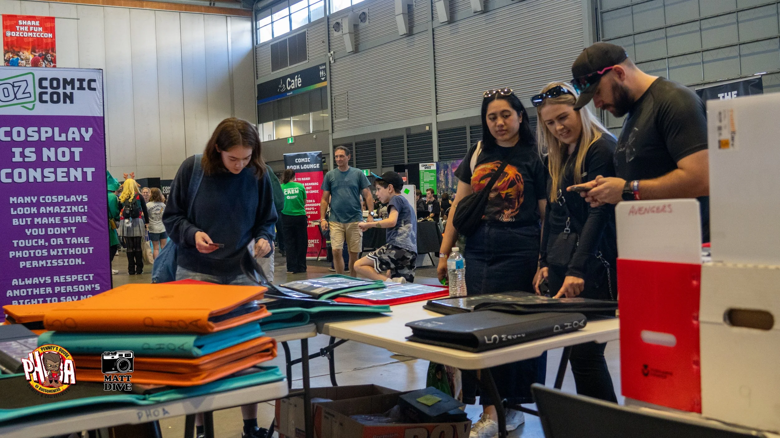 A group of four people at a table with colorful folders and books at a comic convention. A large purple sign with rules about cosplay is on the left. In the background, people walk around and look at booths in a spacious hall.