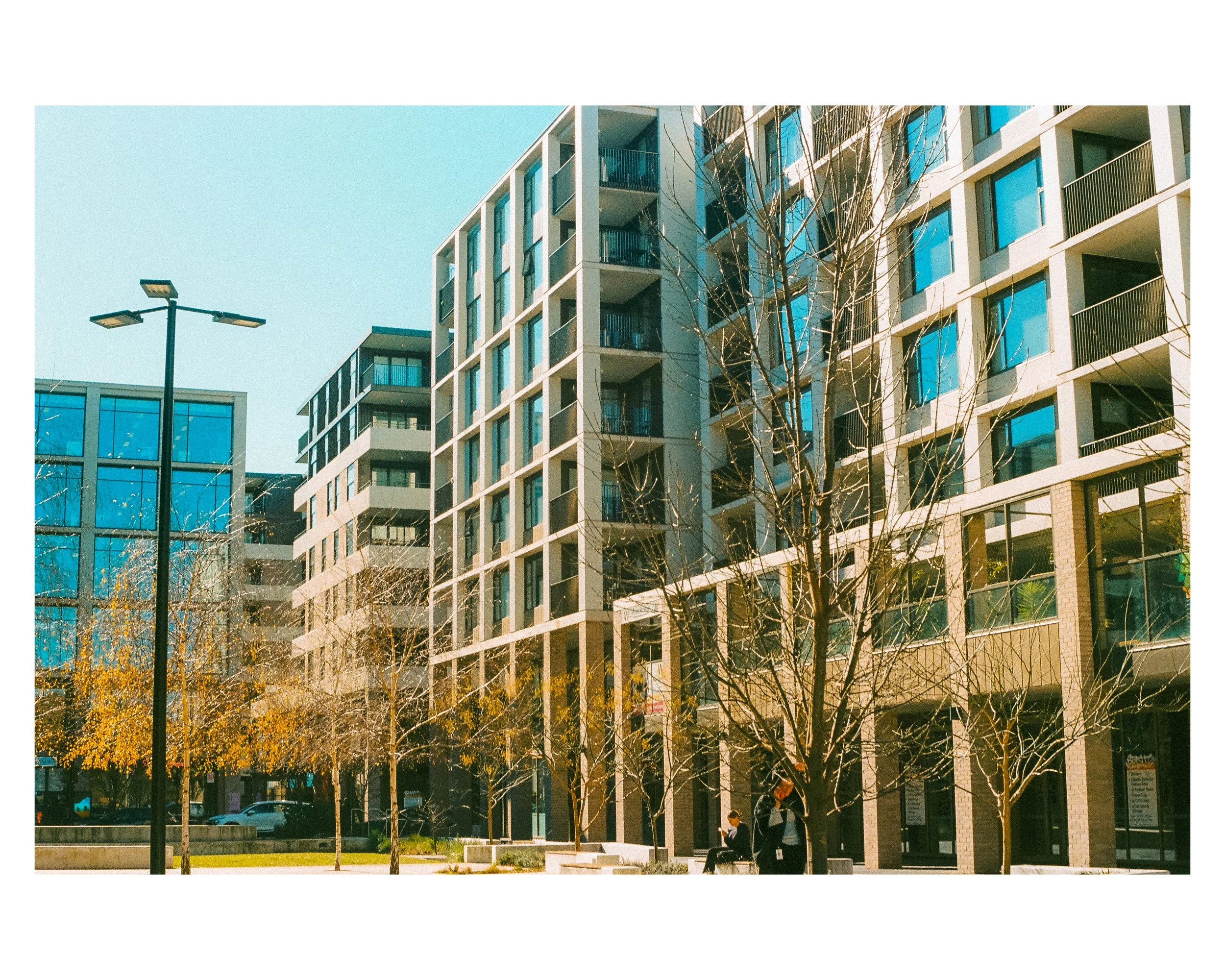 Modern apartment buildings with glass windows and balconies, leafless trees, a park with benches, and people sitting and walking on a sunny day.