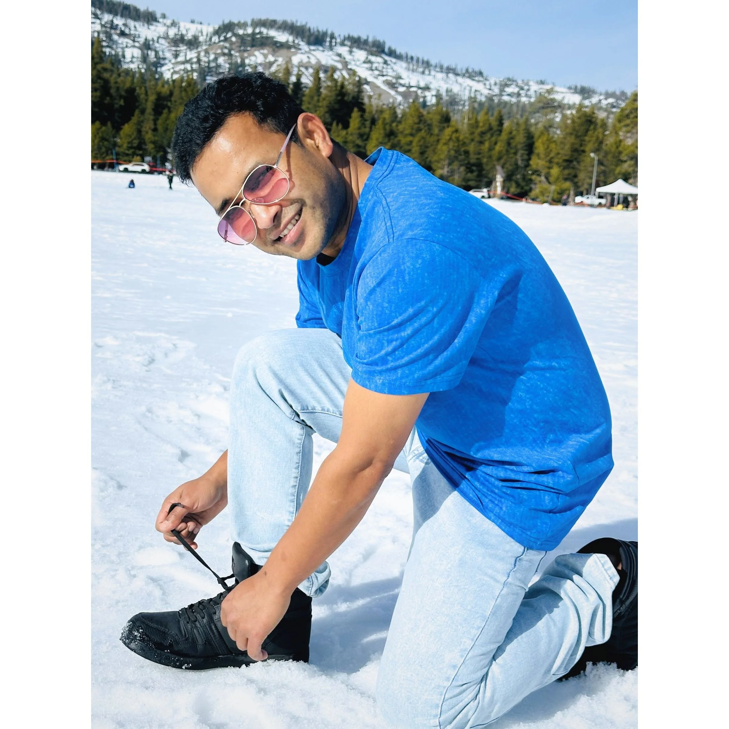 A man in sunglasses and a blue shirt kneeling in the snow and tying his shoelaces, with snow-covered hills and trees in the background.