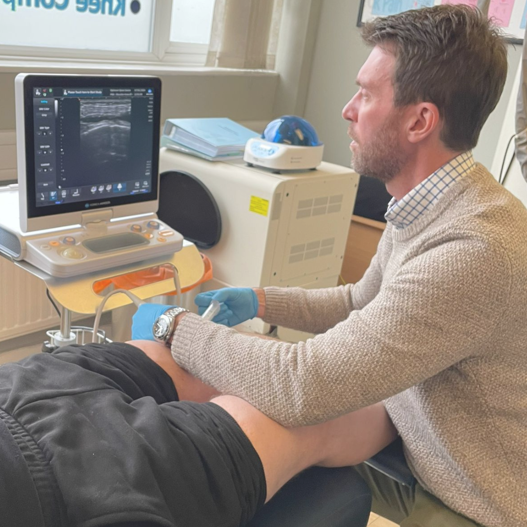 A medical professional in a beige sweater and gloves conducting an ultrasound on a patient's knee, with an ultrasound machine and monitor displaying images in a clinical setting.