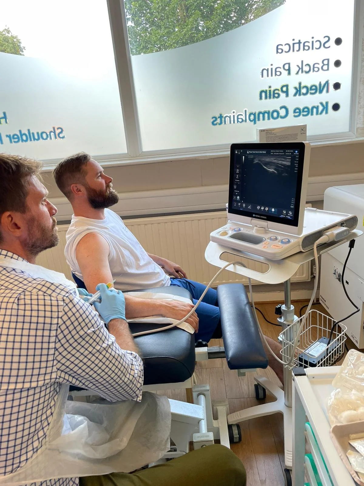 A man is having an ultrasound performed by a medical professional in a clinical setting, with a monitor displaying the ultrasound image.