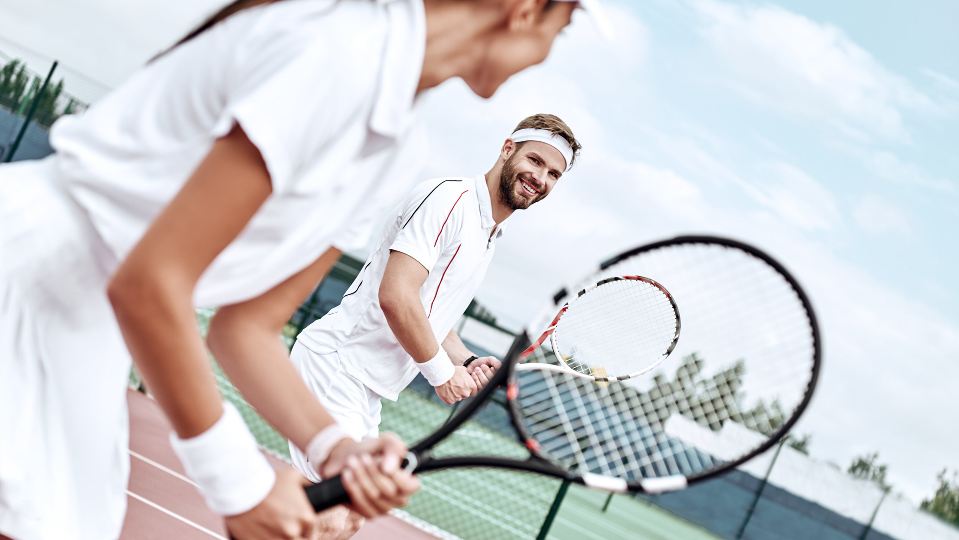 Two people dressed in white tennis outfits holding tennis rackets on a tennis court with a man smiling in the background.