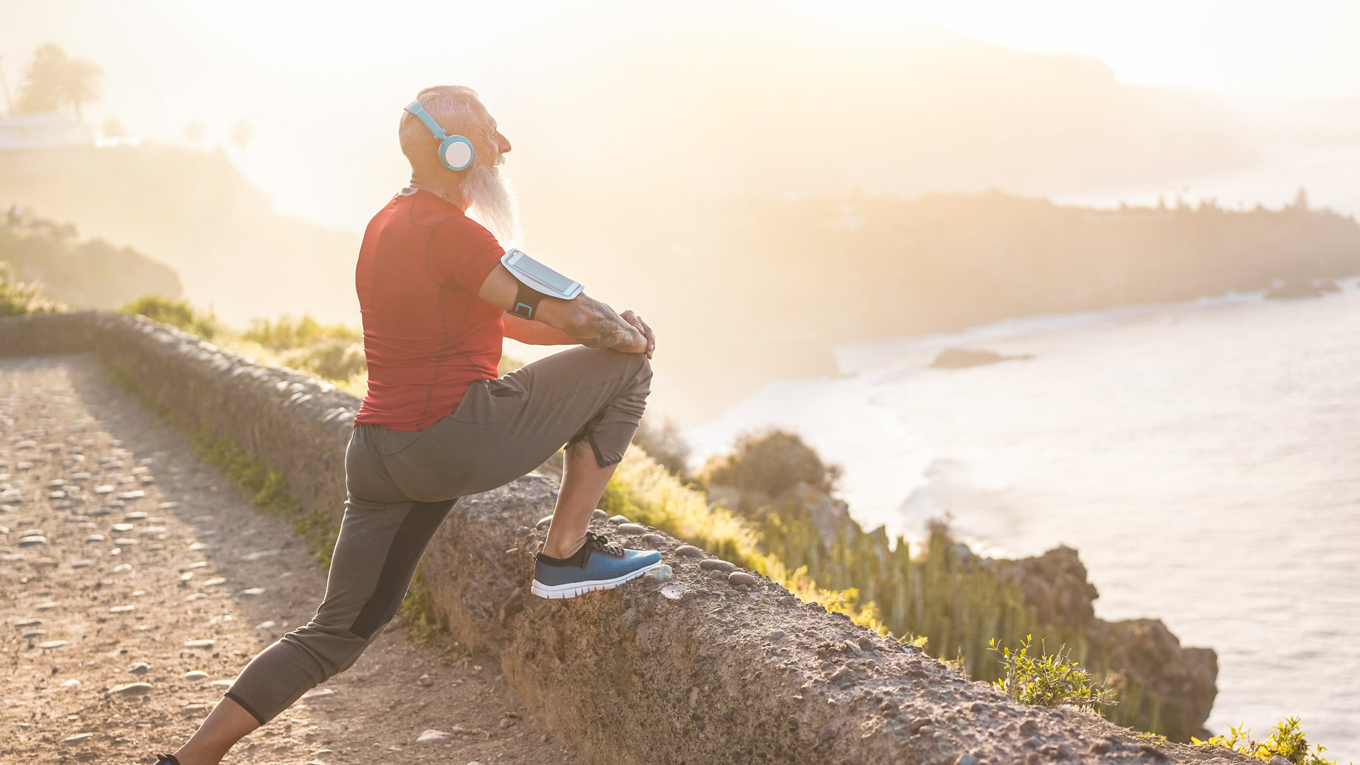 An elderly man with a beard, wearing a red shirt and gray pants, stretching his leg on a concrete barrier during sunrise or sunset along a coastal trail with scenic views of the ocean and cliffs.