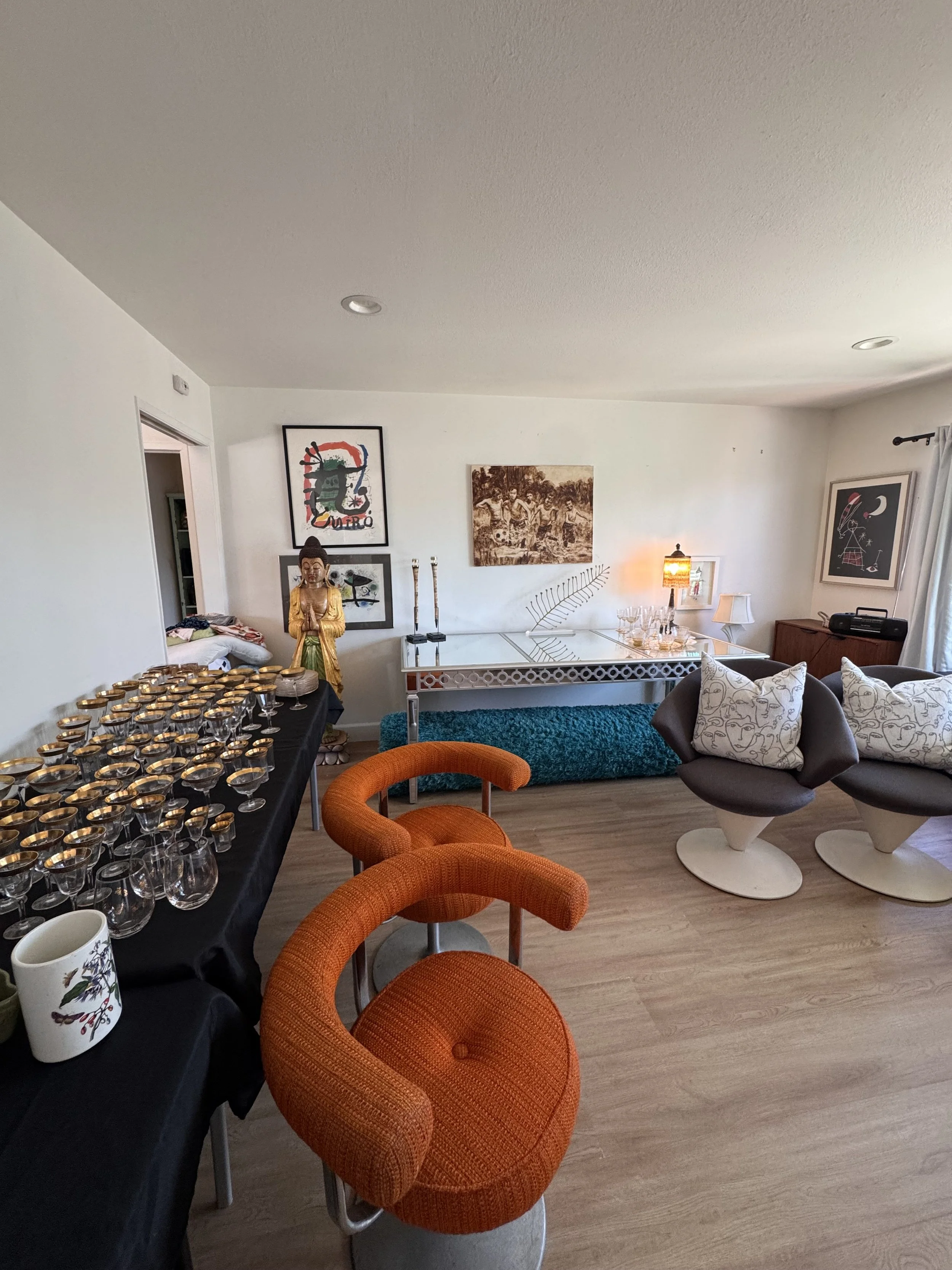 Living room setup with orange upholstered chairs, a black table with tea light candles, various artwork on the walls, and decorative items including a Buddha statue and a table lamp.
