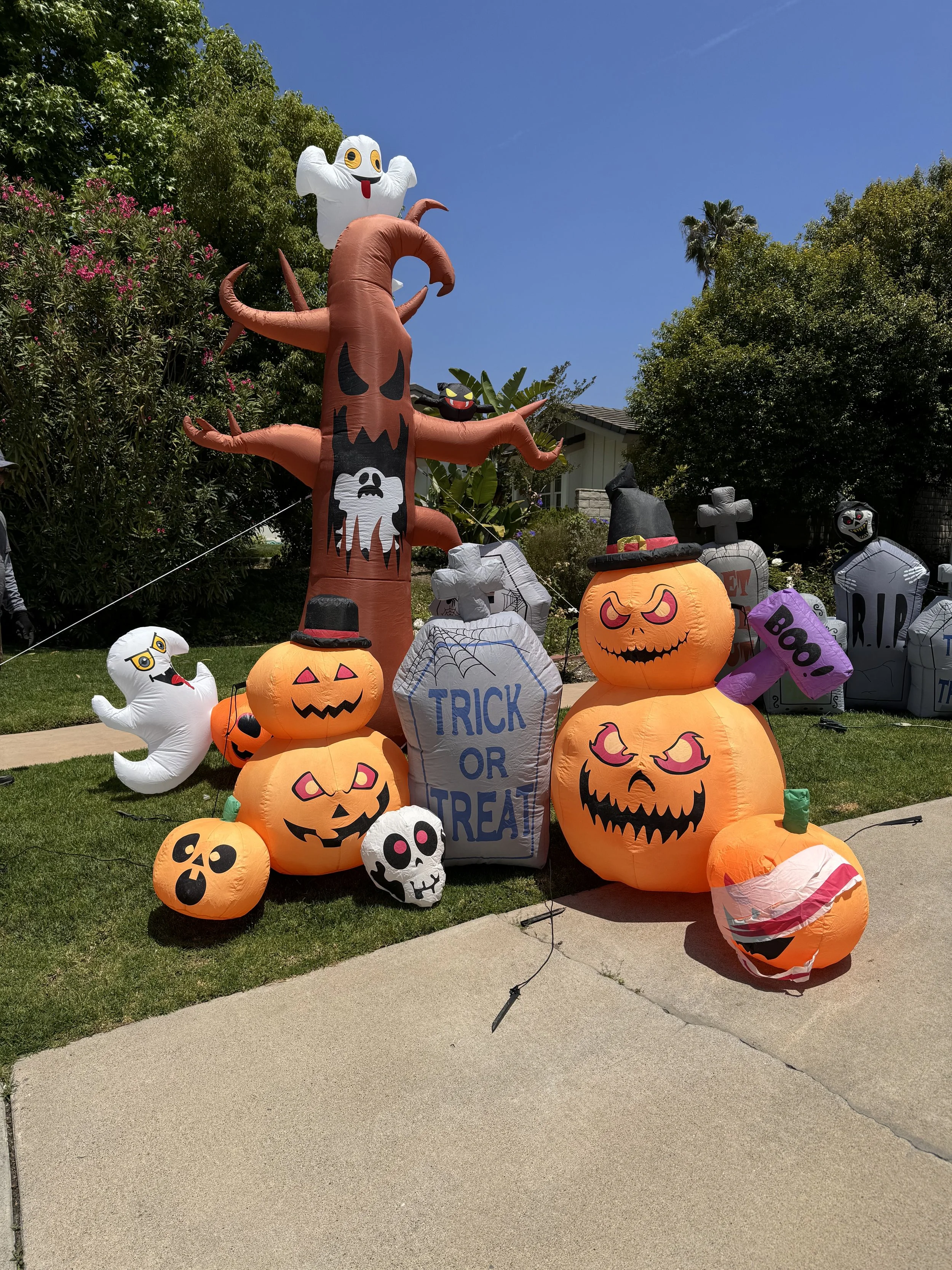 Various inflatable Halloween decorations in a front yard, including a pumpkin with a carved face, a ghost, a black cat with a witch hat, a tombstone that says "Trick or Treat," and other spooky characters, under a sunny blue sky.