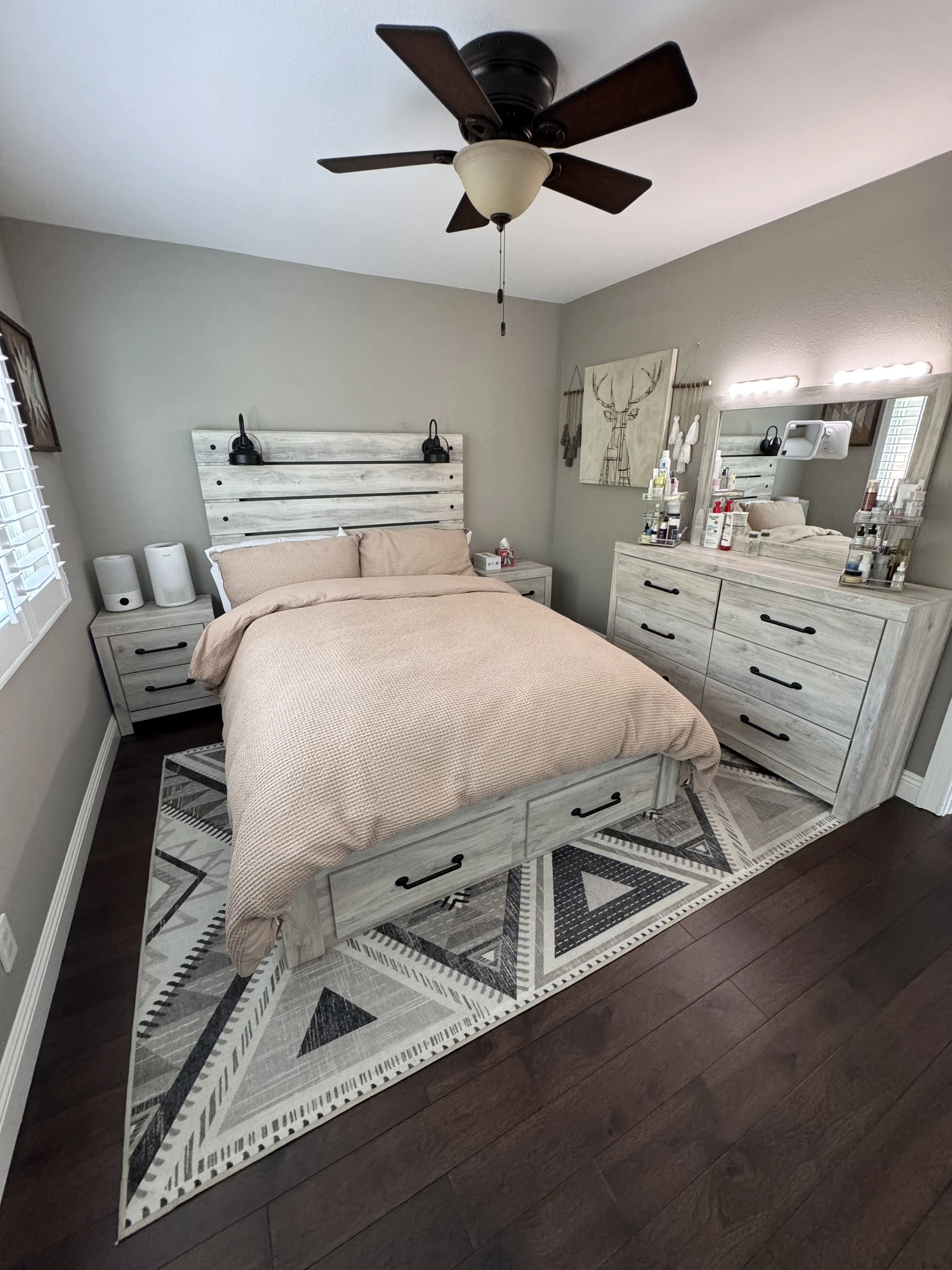 A bedroom with a bed, nightstand, dresser, and mirror. The room has gray walls, a patterned area rug, and a ceiling fan. Items such as tissues, skincare products, and decor are visible.