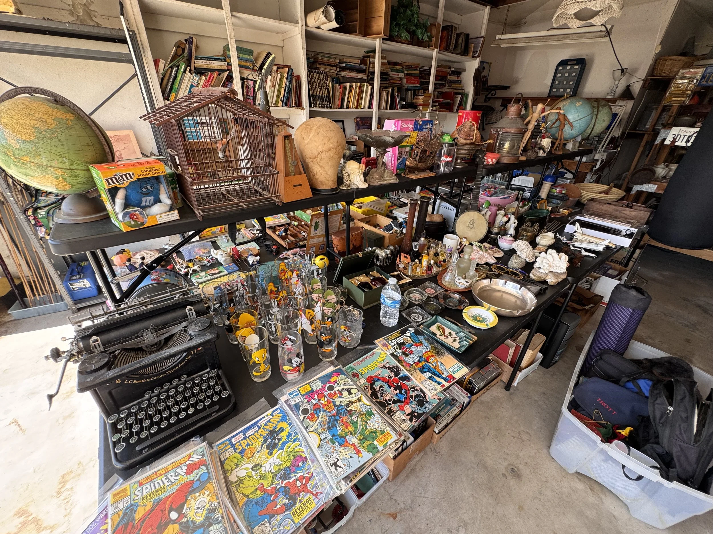 A cluttered space with shelves filled with books, collectibles, and various items. The table in the foreground holds comic books, glasses, and miscellaneous objects, with a vintage typewriter, globes, and small decorative items scattered around. The 