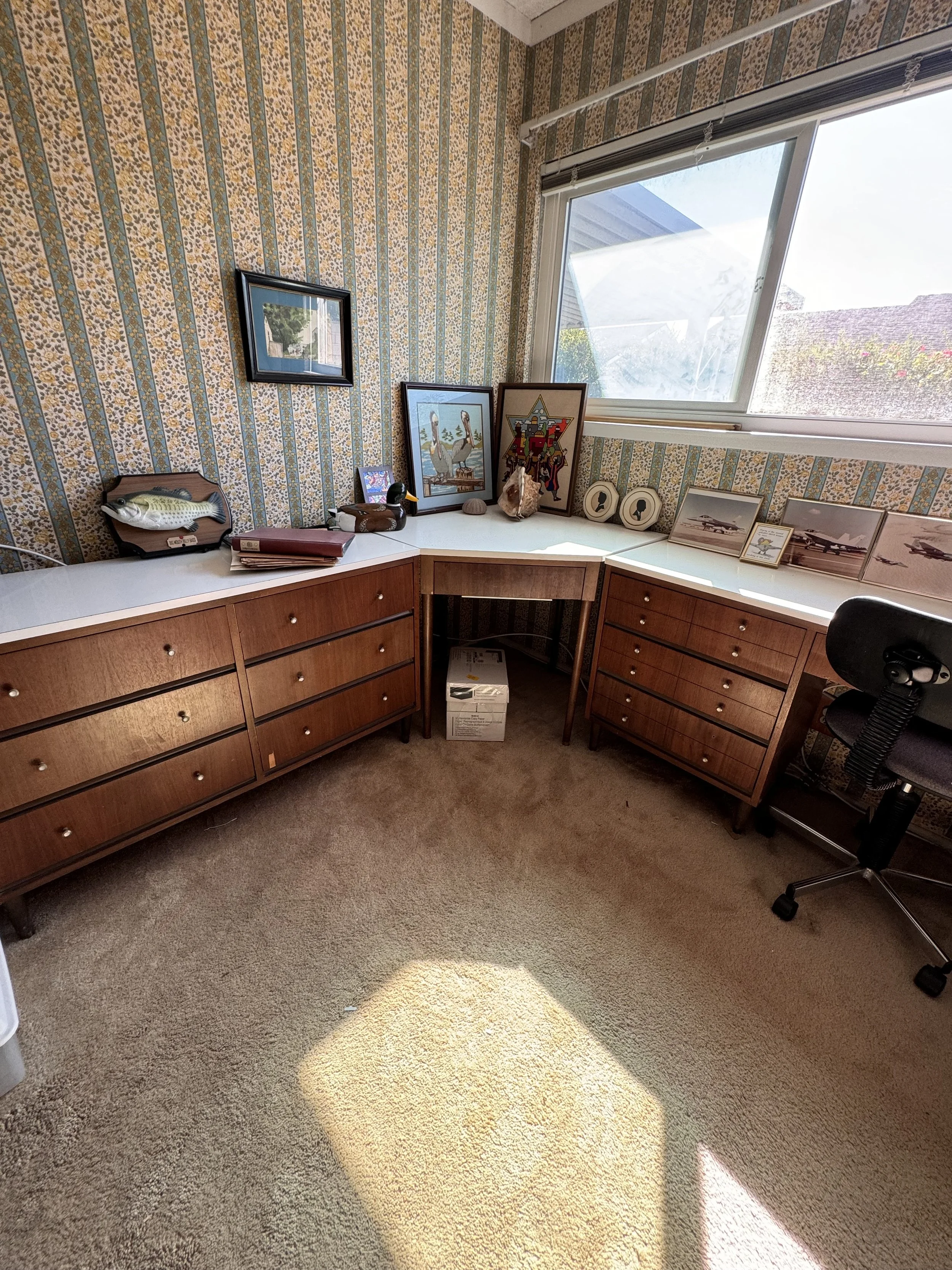 A corner office with mid-century wooden desks with drawers, framed artwork, and decor items on top. A window with a view of a house and garden, and patterned wallpaper.