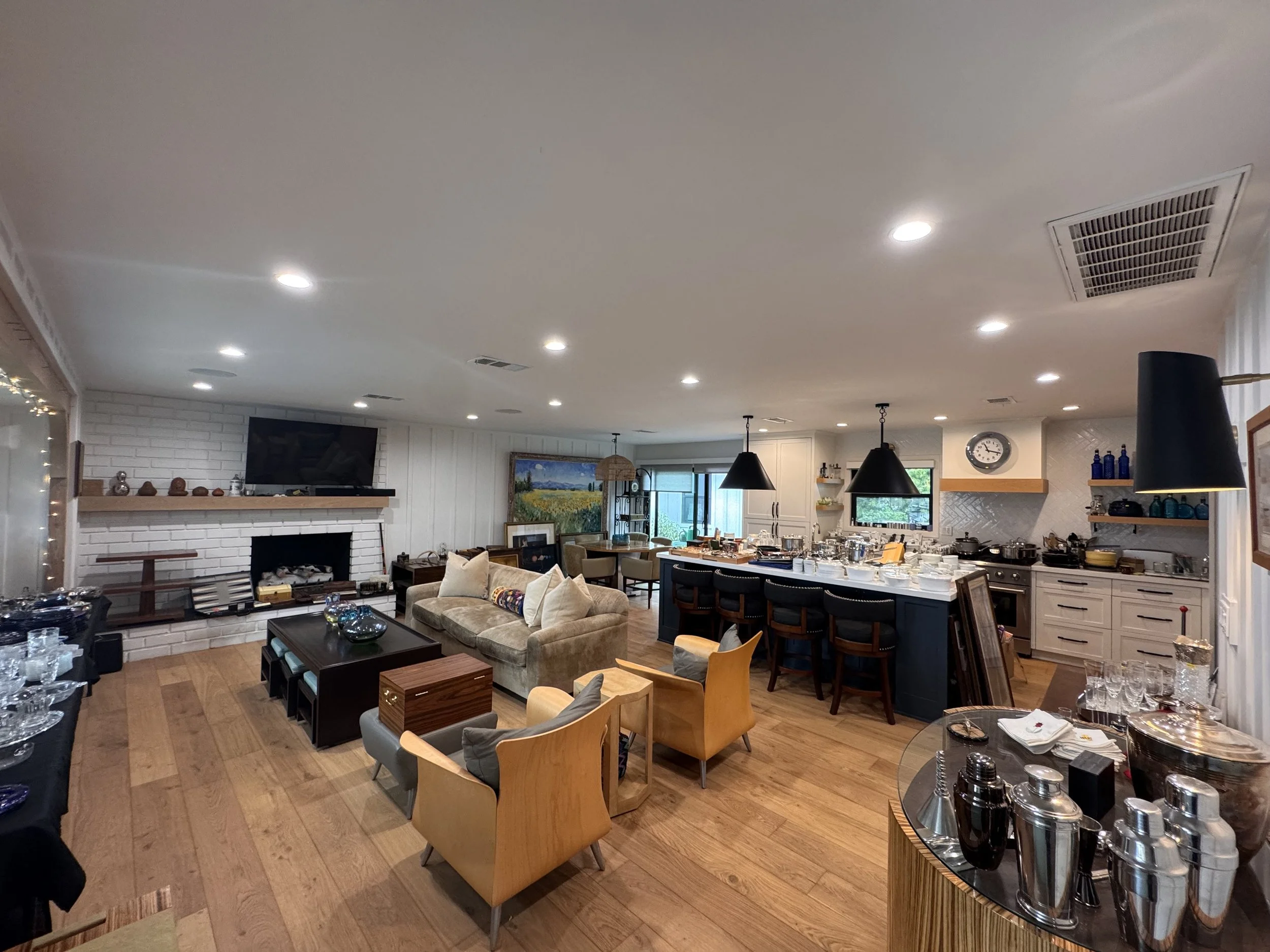 Open-concept living space with a white brick fireplace, flat-screen TV, and dining area. The ceiling has multiple recessed lights and a kitchen island with barstools. There are various decorations, artwork, and kitchenware throughout the room.