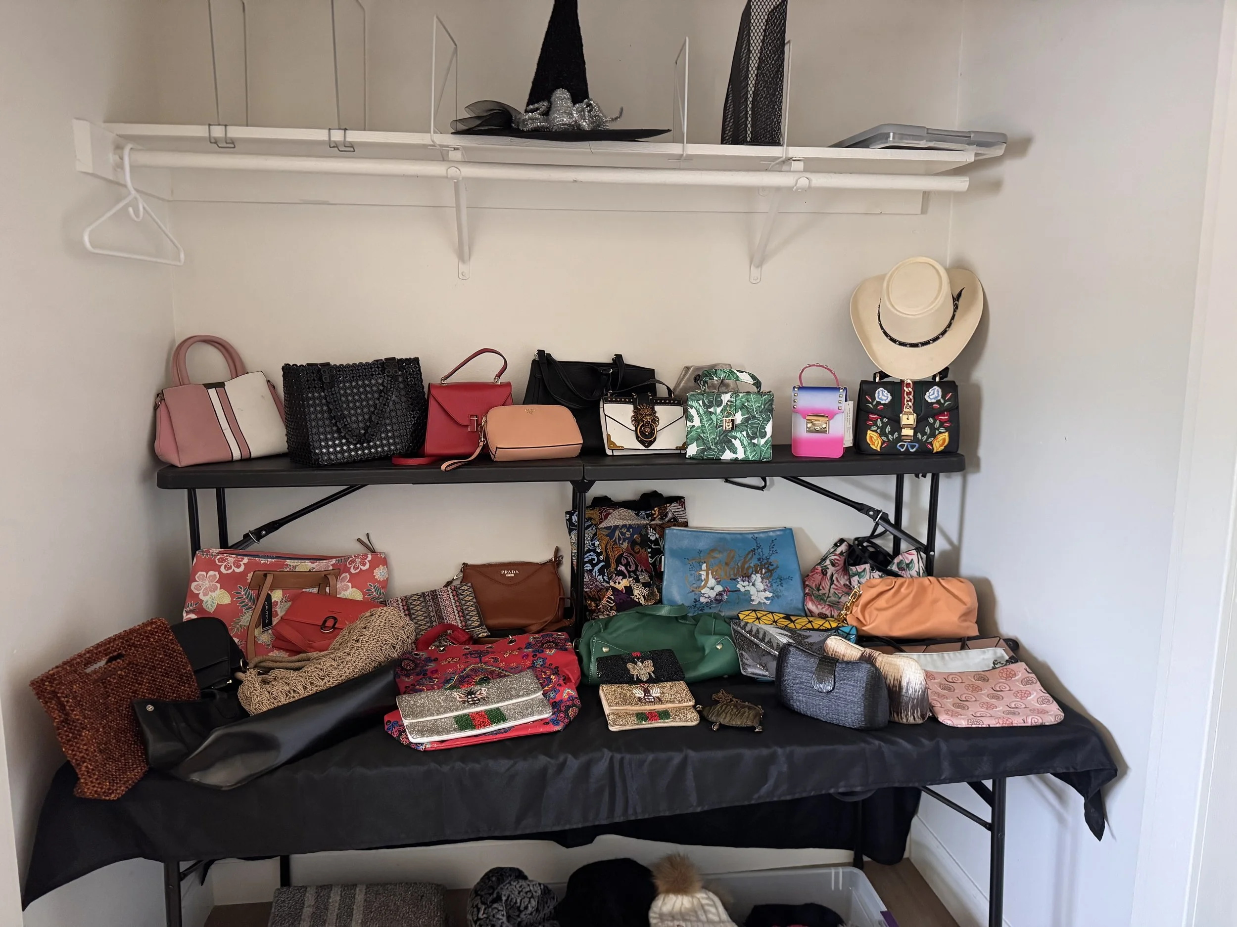 A table and shelves filled with various handbags and purses, along with some hats and accessories, in a room with cream-colored walls.