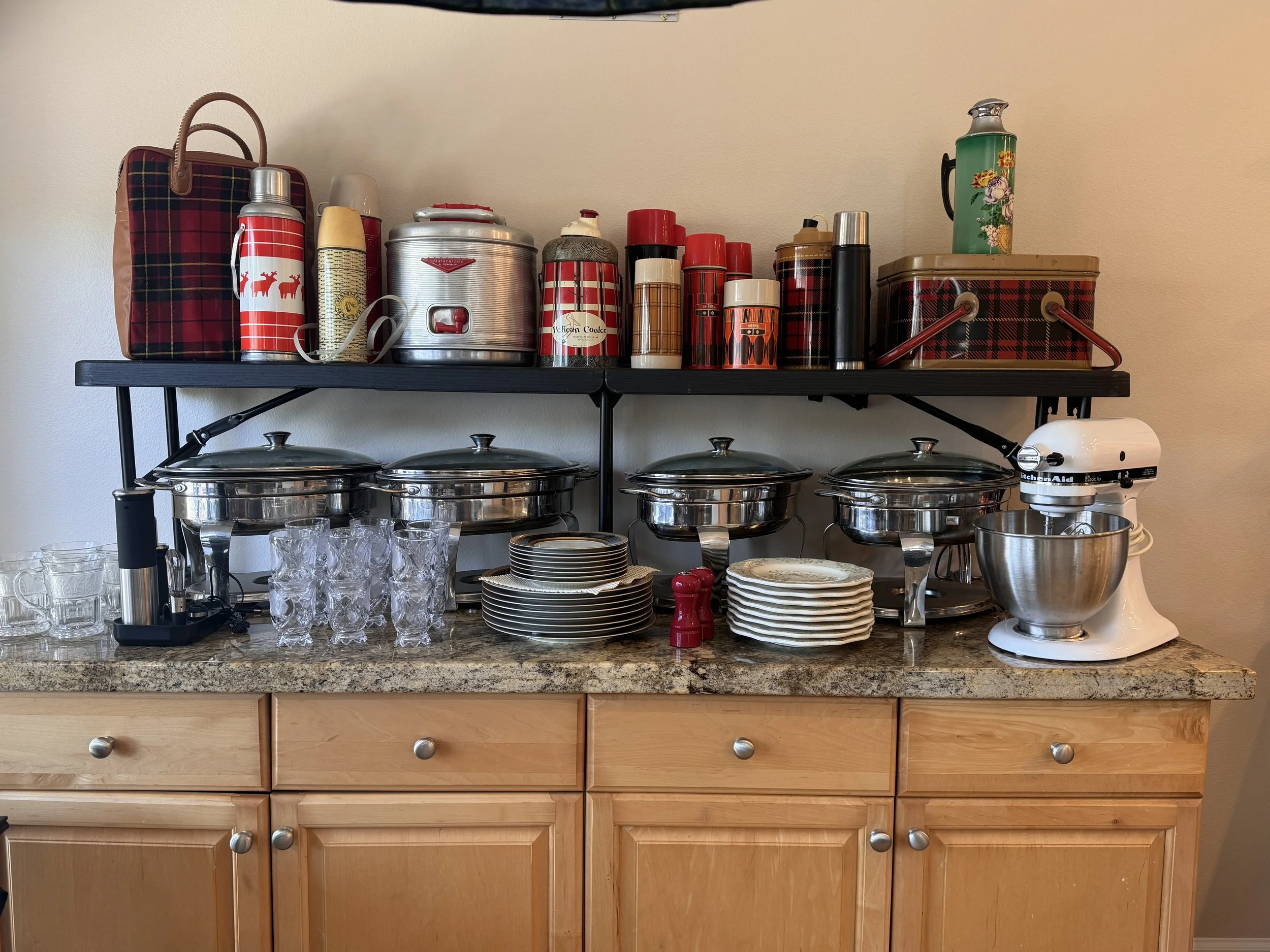 A kitchen counter with glassware, plates, and a stand mixer, above which a black shelf holds kitchen themed containers, thermoses, and cookware with a matching plaid pattern and a green mug with flowers.