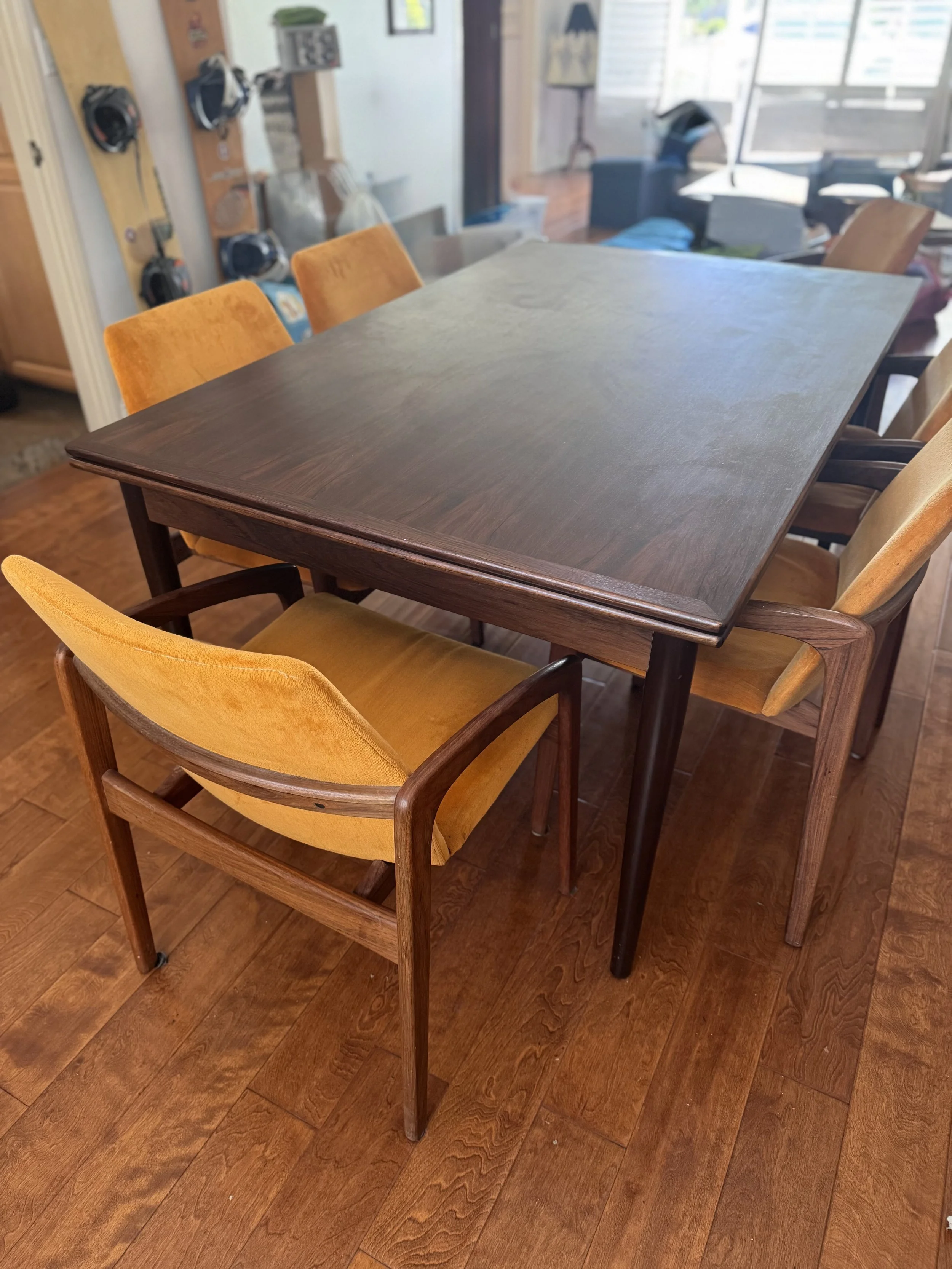 A wooden dining table surrounded by six yellow upholstered chairs in a cozy kitchen or dining area, with a hardwood floor and sunlight coming through large windows.