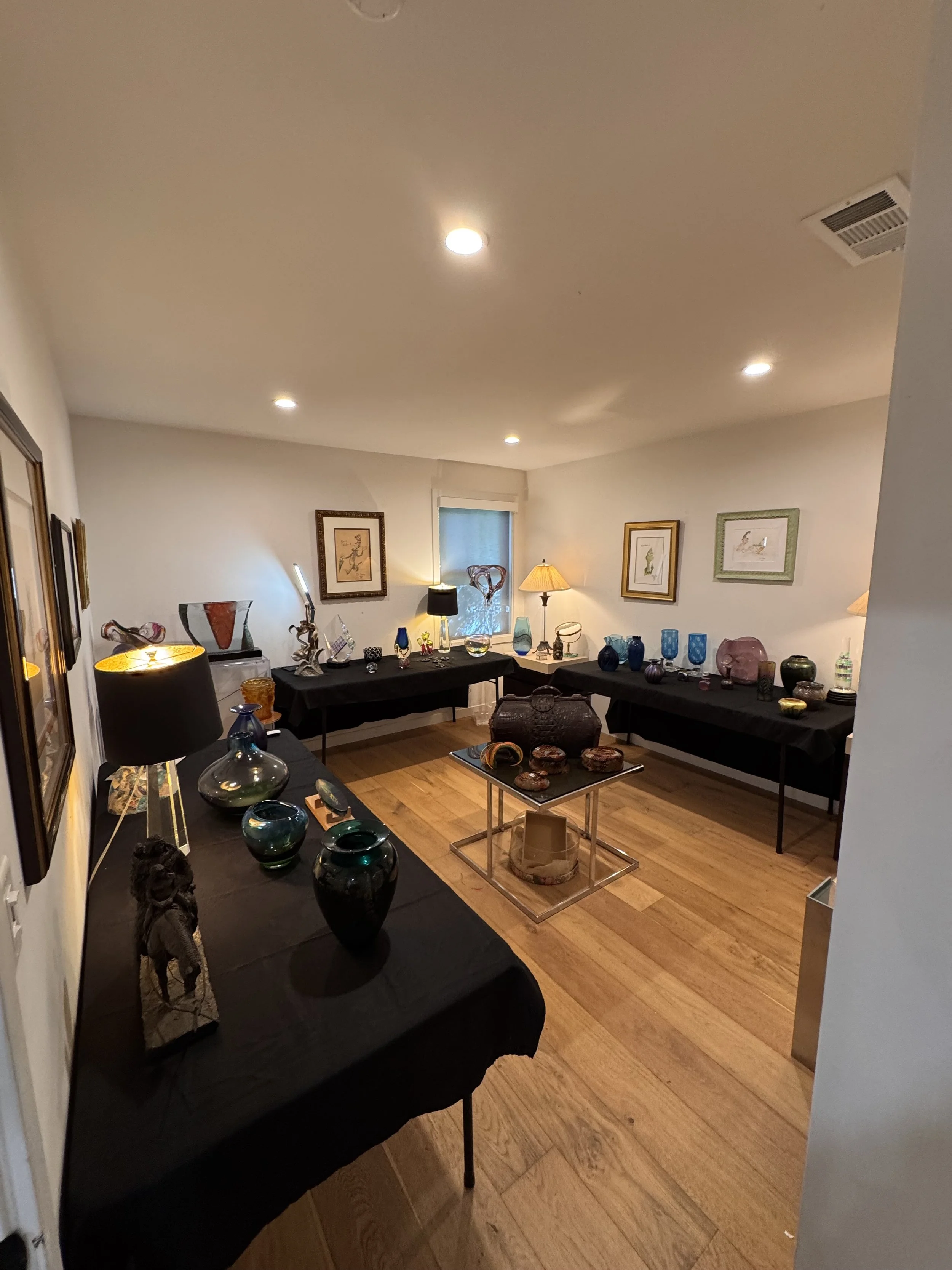 A room with wooden flooring and white walls decorated with framed artwork. Several tables are covered with black cloths, displaying various glass and ceramic items, including vases, bowls, and sculptures. There are lamps providing warm lighting, and 