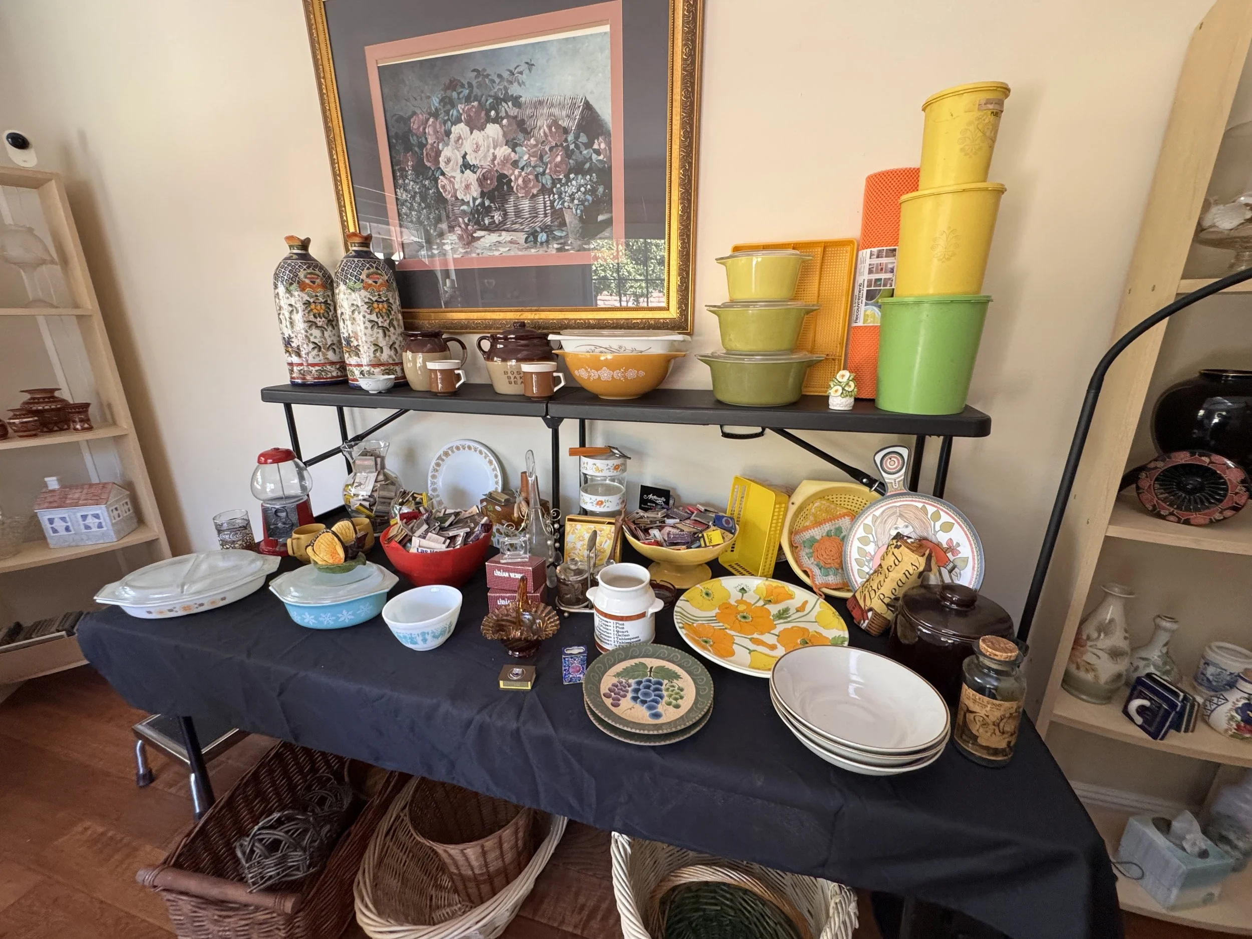 Table filled with various dishes and kitchenware, with a shelf of decorative bottles and framed artwork in the background.