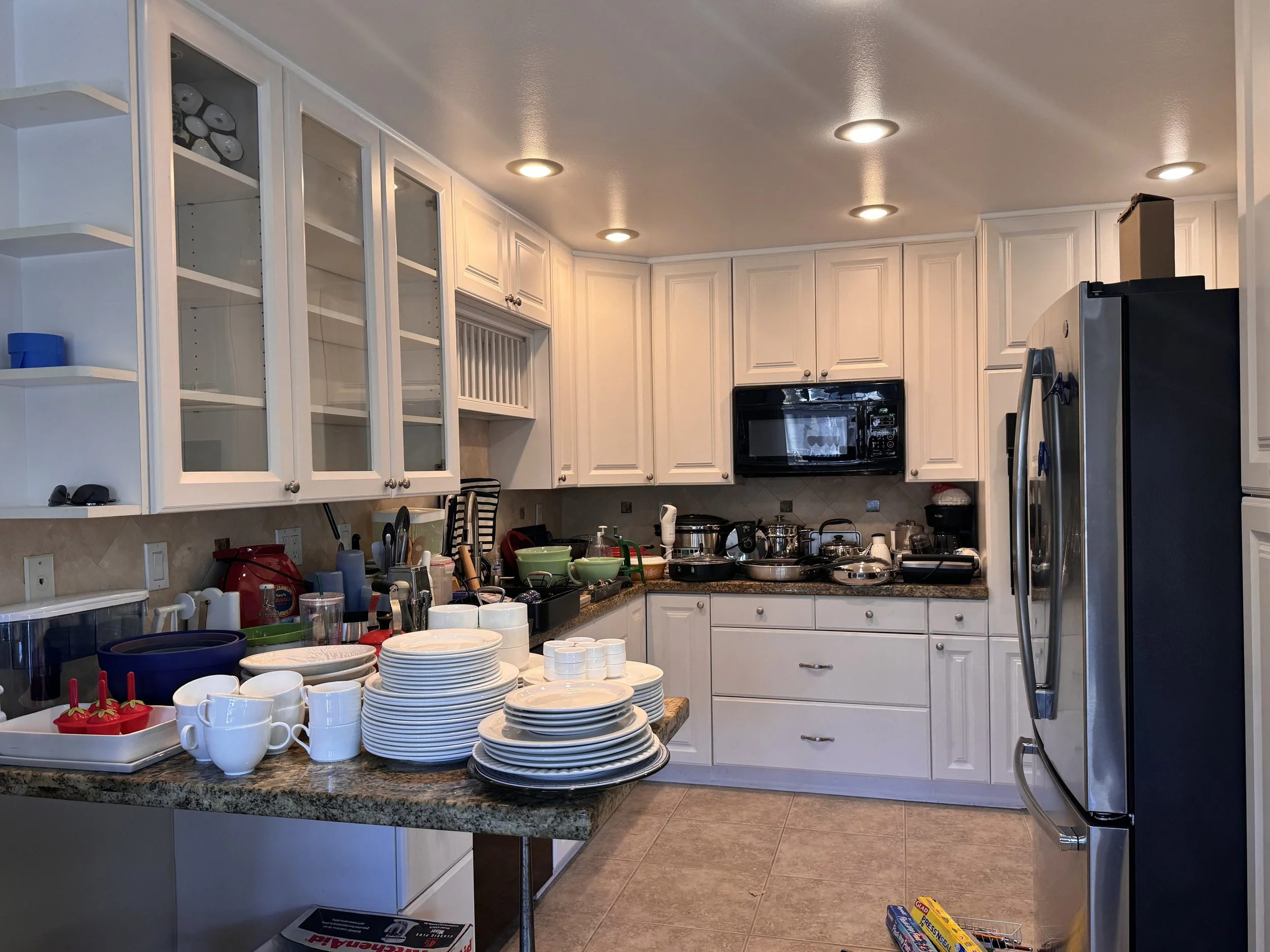 Messy kitchen with dishes, cups, and utensils on the countertop. Open shelves, microwave, refrigerator, and kitchen cabinets are visible. Counter clutter includes food containers and kitchen tools.