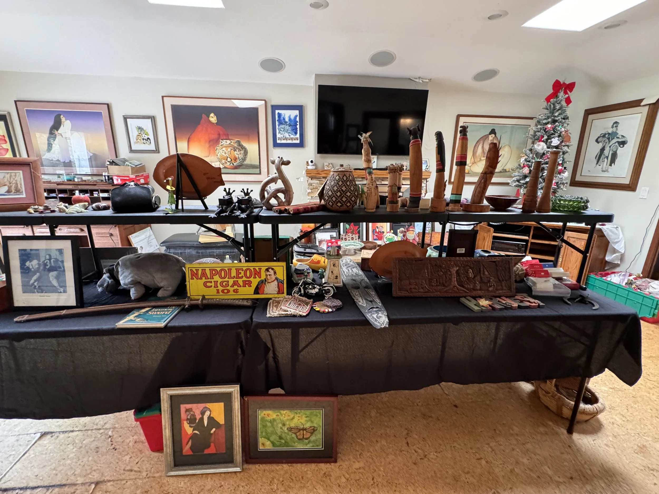 A table displays vintage and collectible items, including framed artwork, small sculptures, a miniature motorcycle, decorative vases, and a sign reading 'Napoleon Cigar 10¢'. In the background, framed paintings and a small Christmas tree with red bow