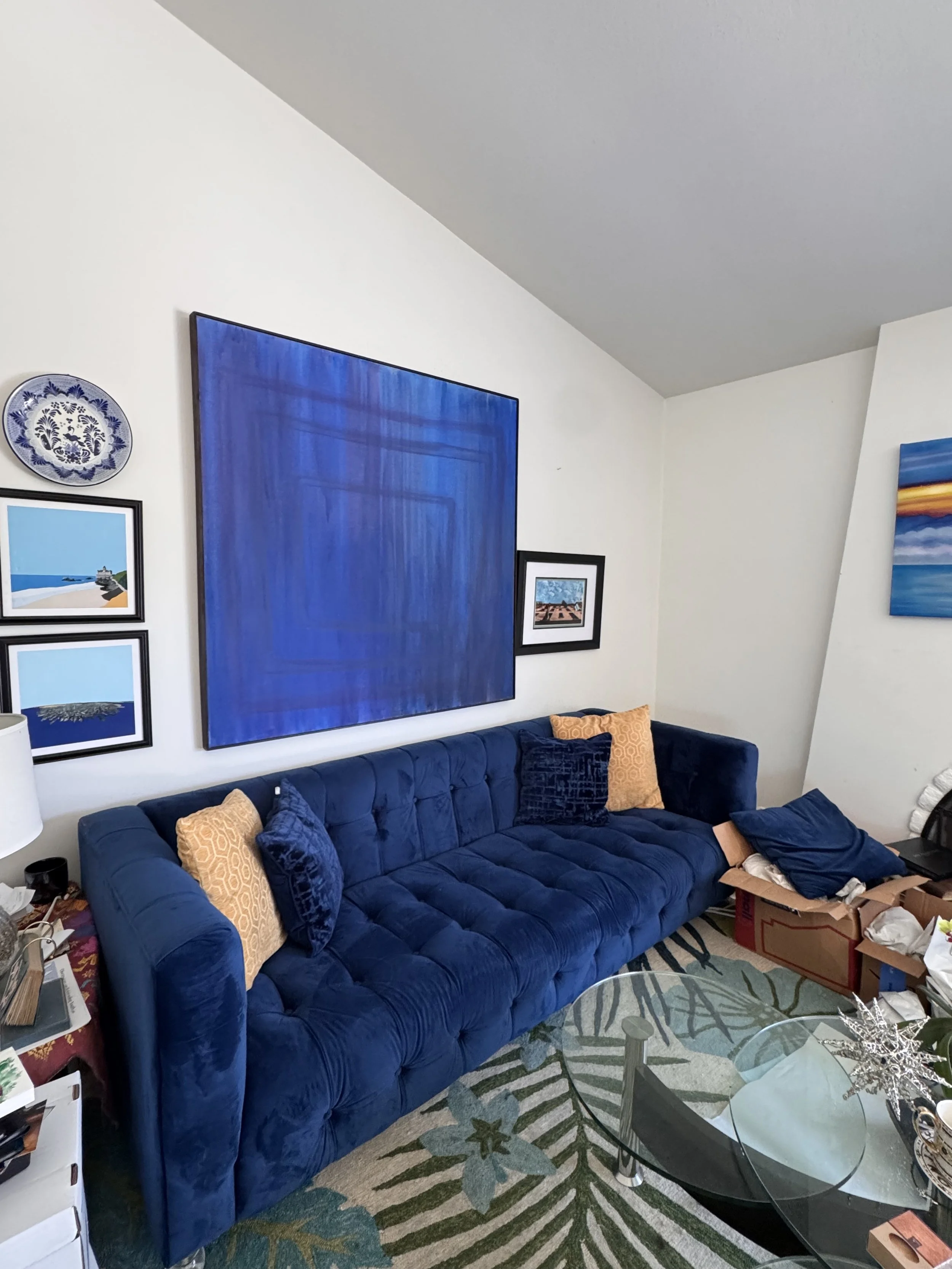 Living room with a blue upholstered sofa with yellow and blue pillows, wall art, a glass coffee table, a patterned rug, and clutter including a cardboard box, a stack of papers, and personal items.