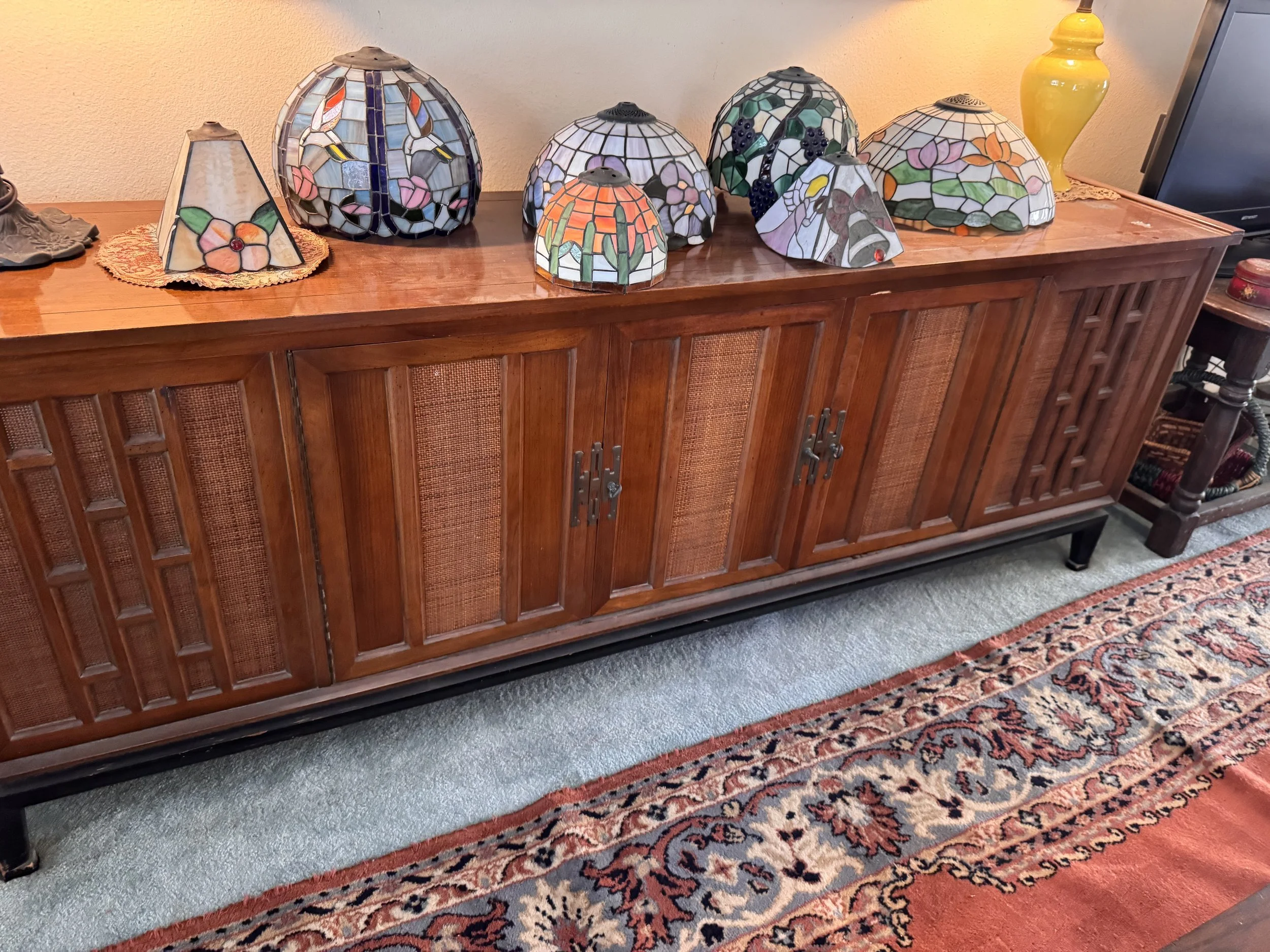 Collection of stained glass lamps with floral and animal designs on a wooden sideboard, next to a yellow vase and a computer monitor, placed on a patterned rug.