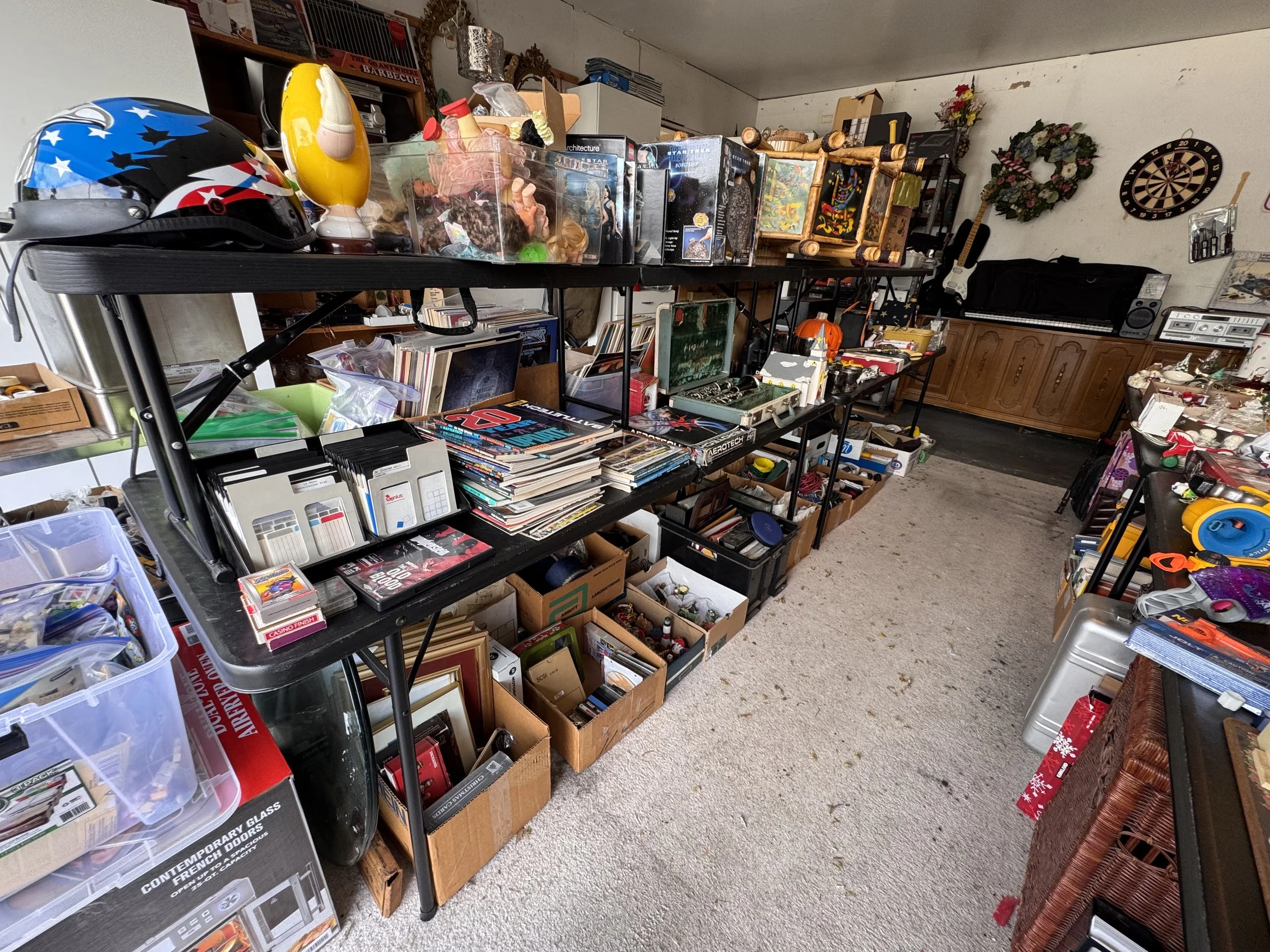 A room filled with various items on black shelving units, including helmets, dolls, books, and collectibles, with a dartboard and a guitar on the wall.