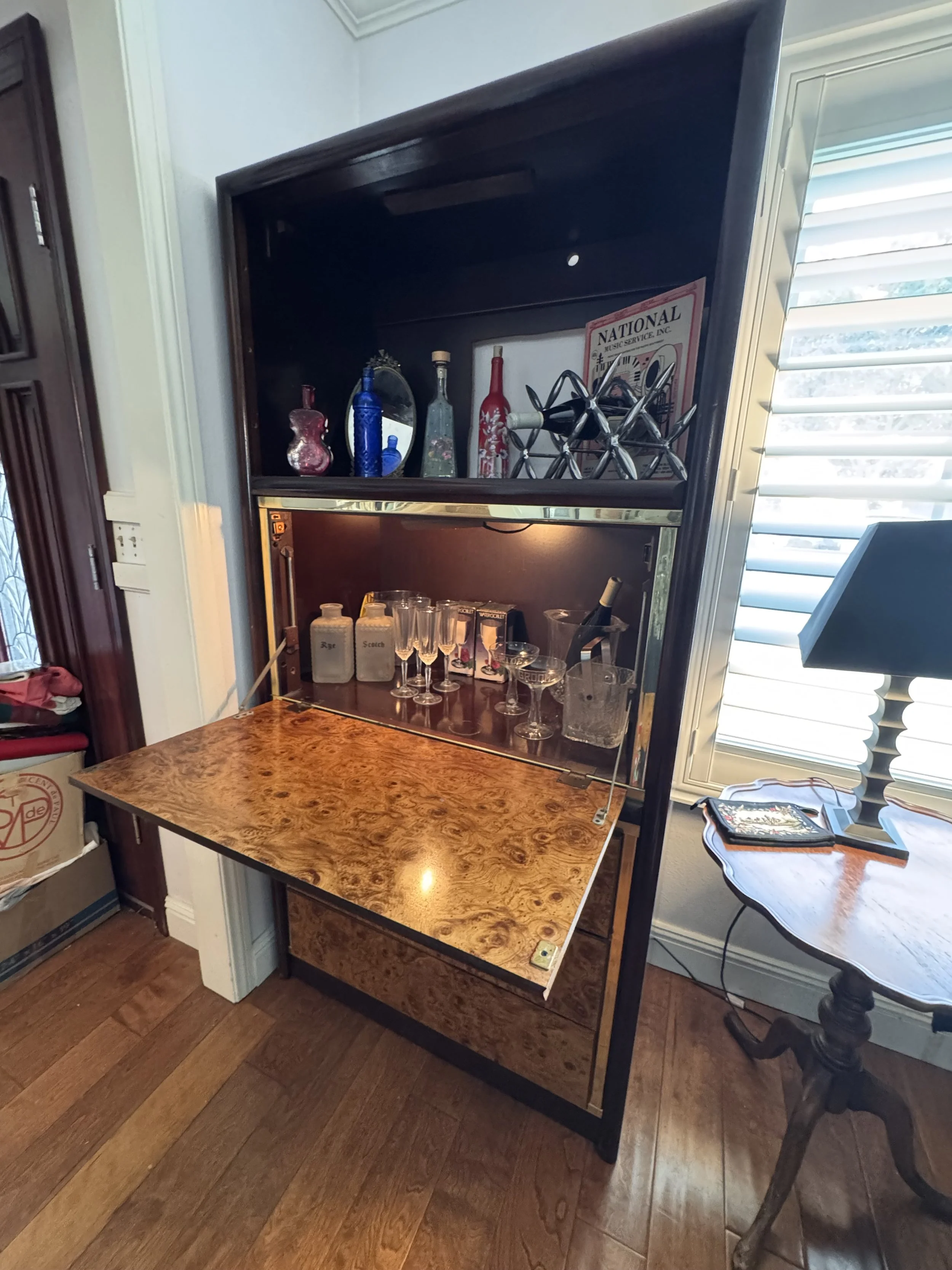 A vintage style bar cabinet or liquor display with a wood and black frame, open front, and a built-in shelf. The shelf holds liquor bottles, glasses, a mirror, and decorative items. The cabinet is placed next to a window with white blinds, on a hardw