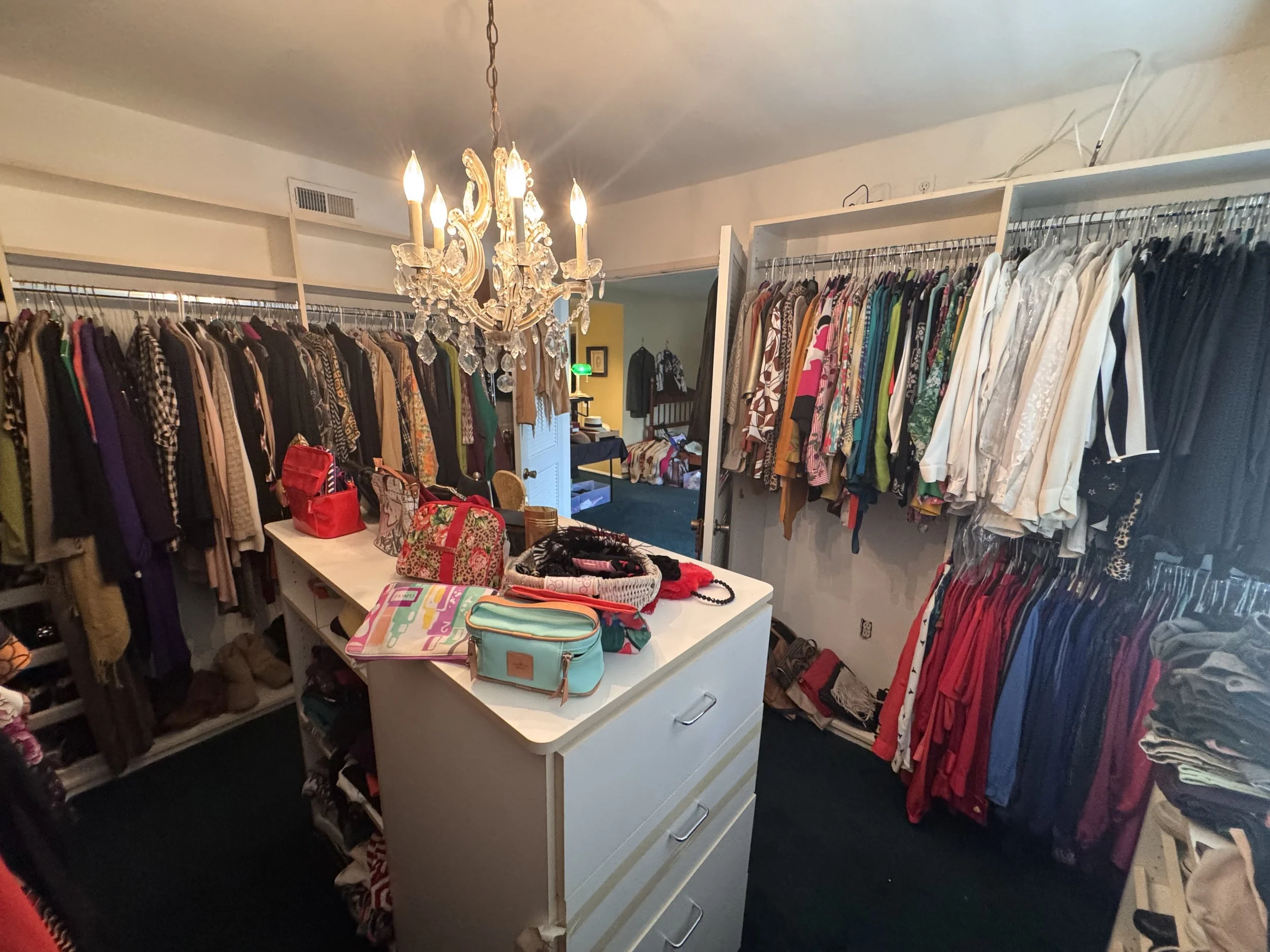 A walk-in closet with clothing racks filled with various garments on both sides and a white dresser in the center holding bags and accessories. A chandelier hangs from the ceiling, illuminating the space.