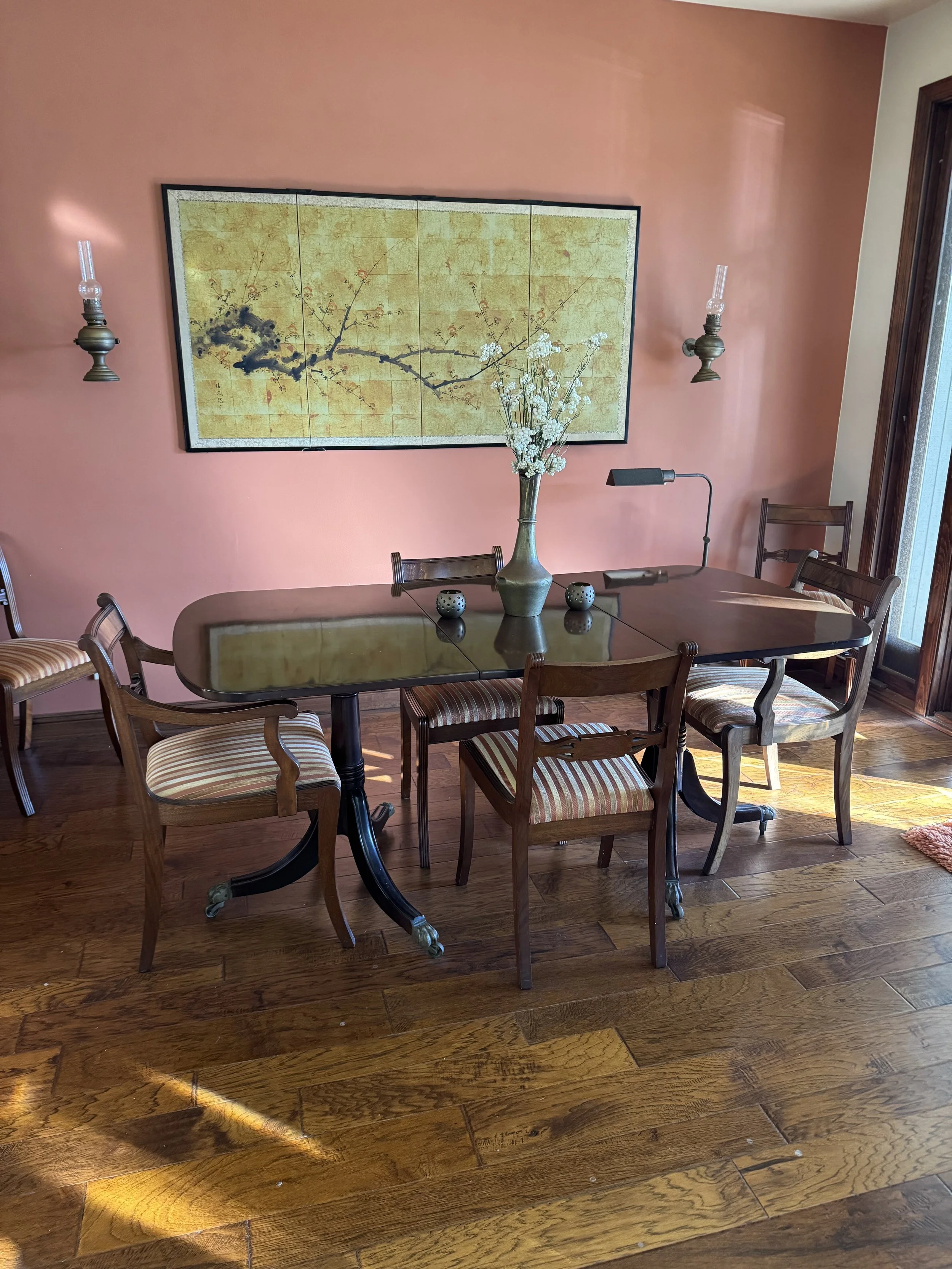 A dining room with a wooden table and six chairs, a pink wall with Asian style art, and decor including a vase with white flowers, two small candles, and a wall-mounted lamp.
