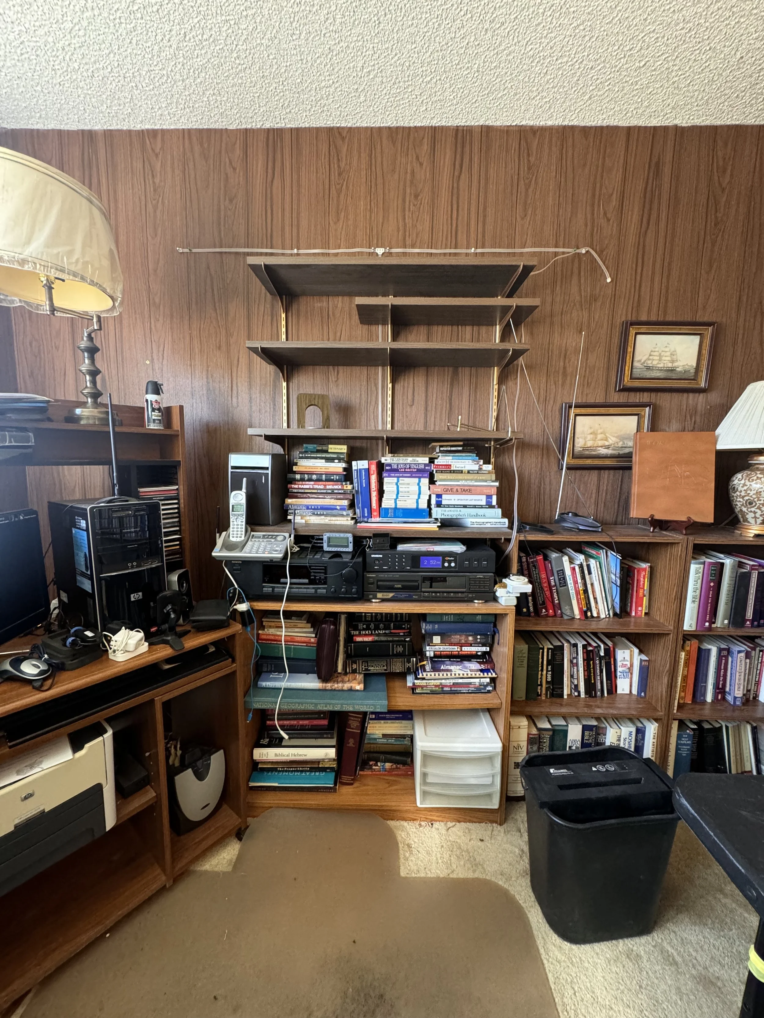 A cluttered home office with a wooden wall, bookshelves filled with books, a desk with electronic devices, and a large black trash bin.