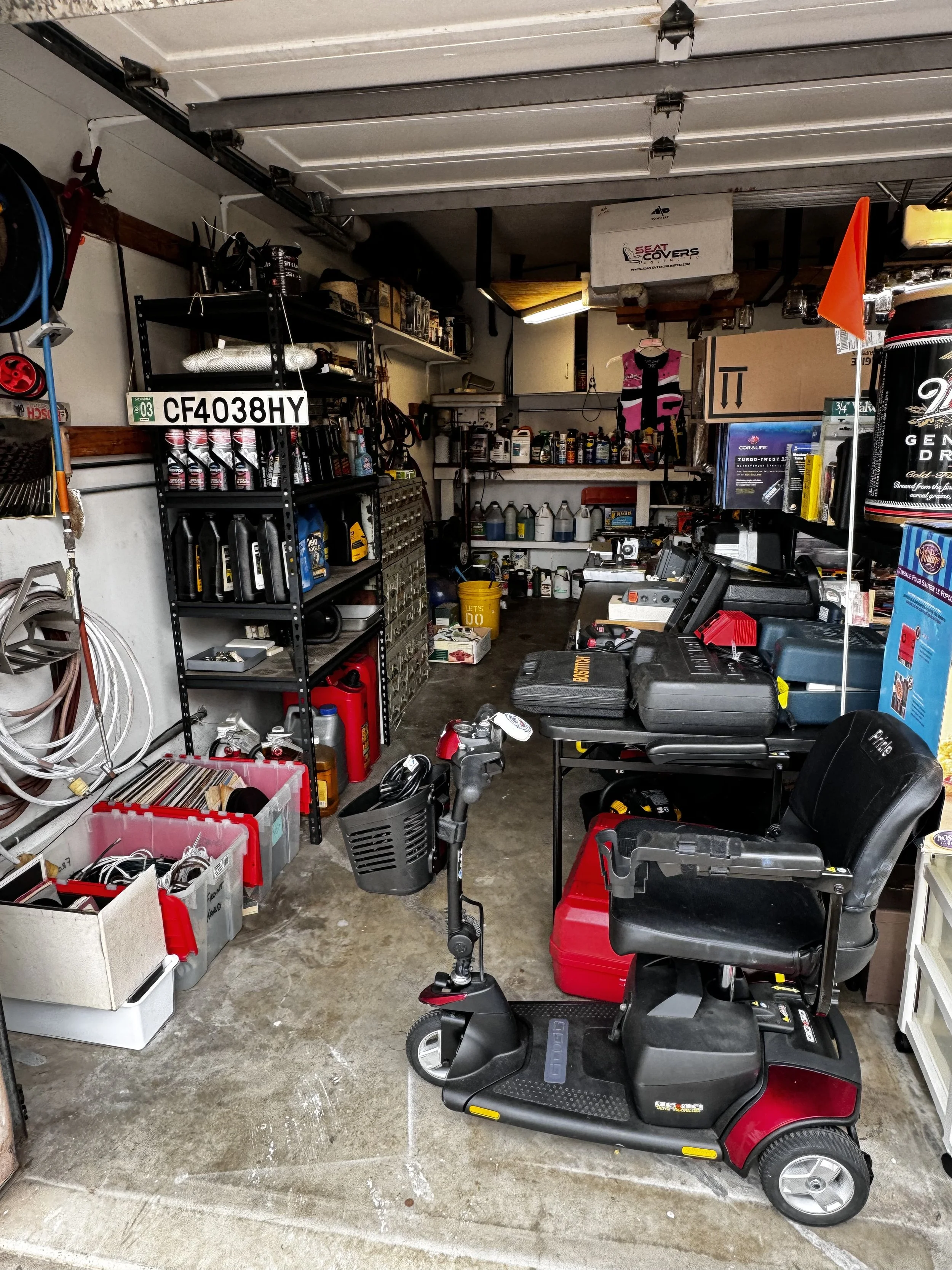 A garage filled with various tools, shelves with motor oil bottles, a robotic lawn mower in the center, and storage boxes, including a helmet and gardening tools hanging on the walls.