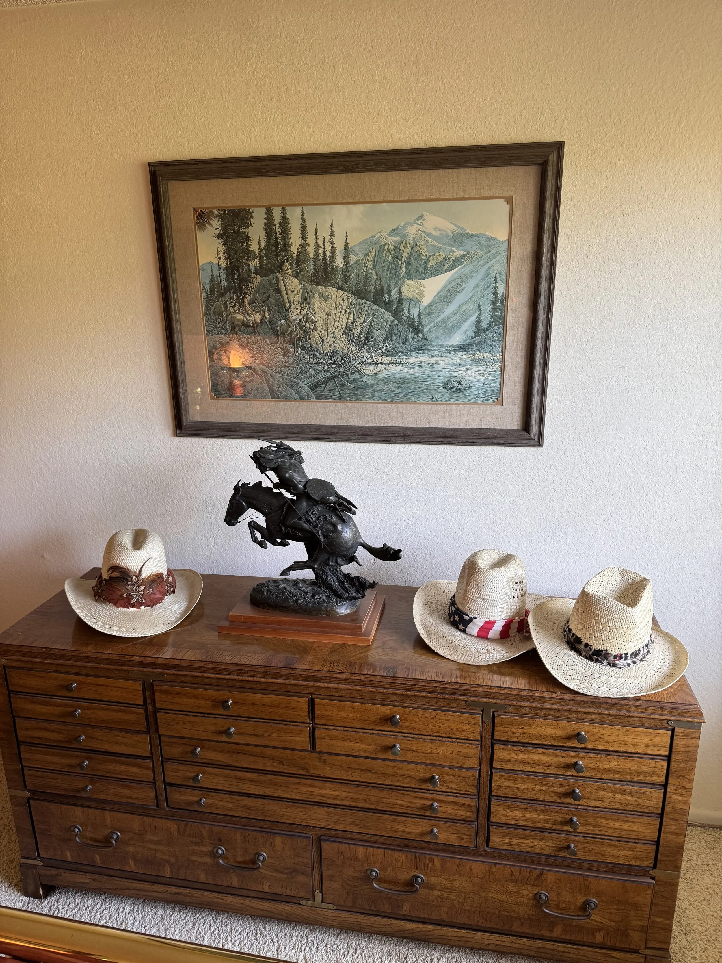 A wooden dresser with multiple drawers featuring a bronze horse sculpture and three straw hats with different band designs on top, against a white wall with a framed mountain landscape painting.