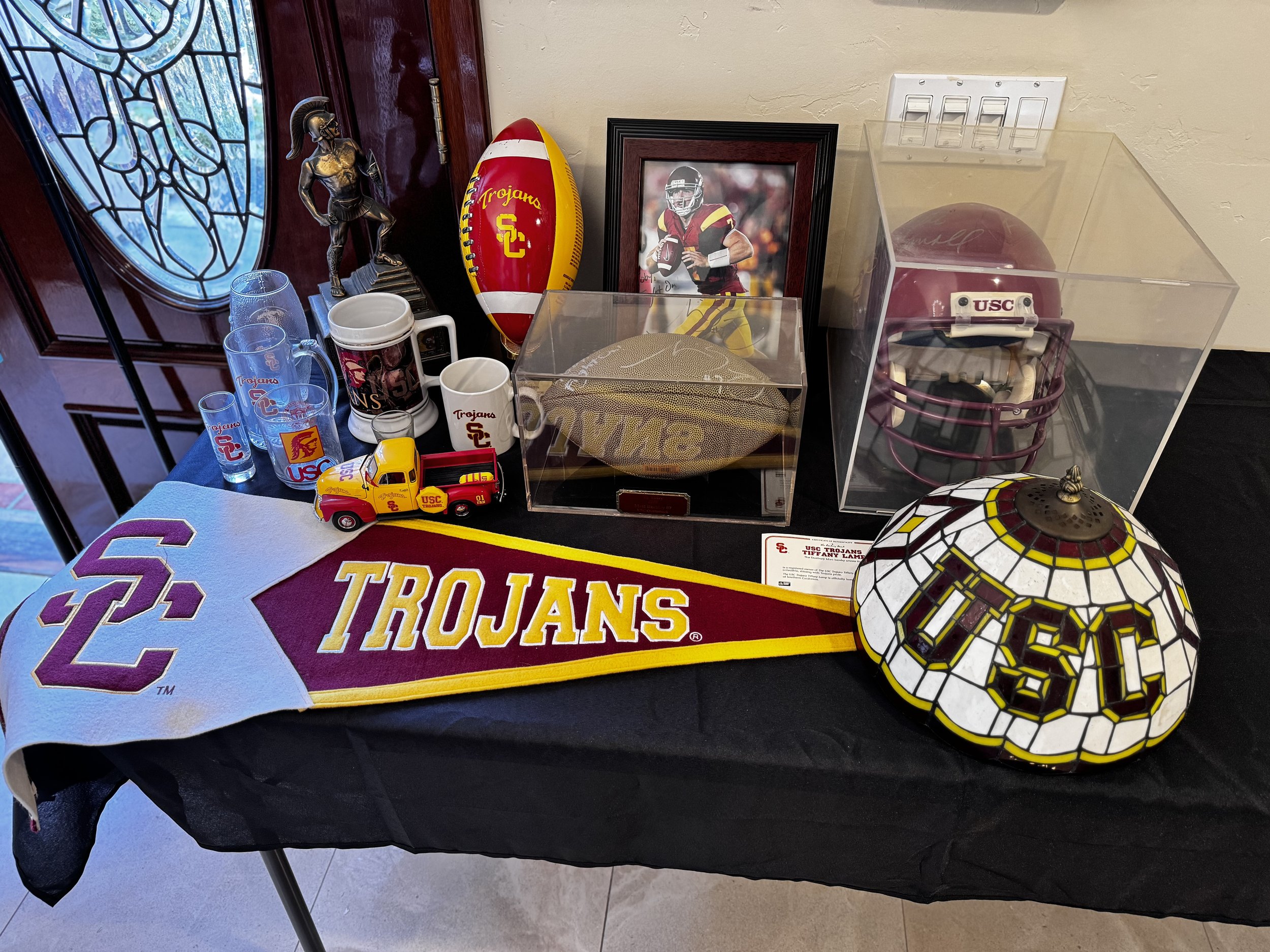 Display of University of Southern California Trojans memorabilia, including a USC football, framed photo of a football player, Trojan helmet, small model fire truck, and USC-branded glassware, arranged on a table with a black tablecloth.