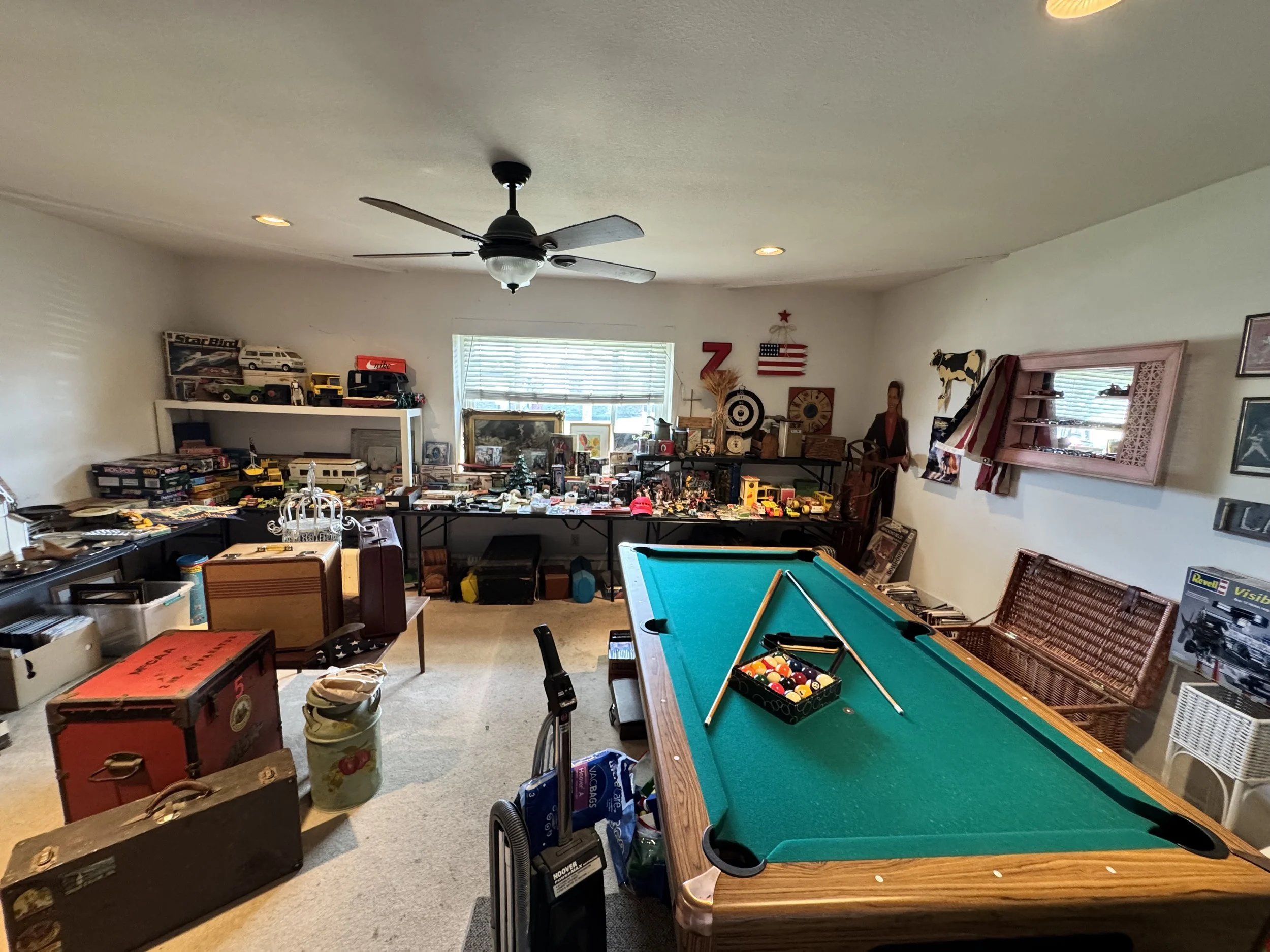 A cluttered basement or game room filled with various toys, collectibles, and furniture, including a pool table, shelves with model cars, and a cardboard box with vintage items.