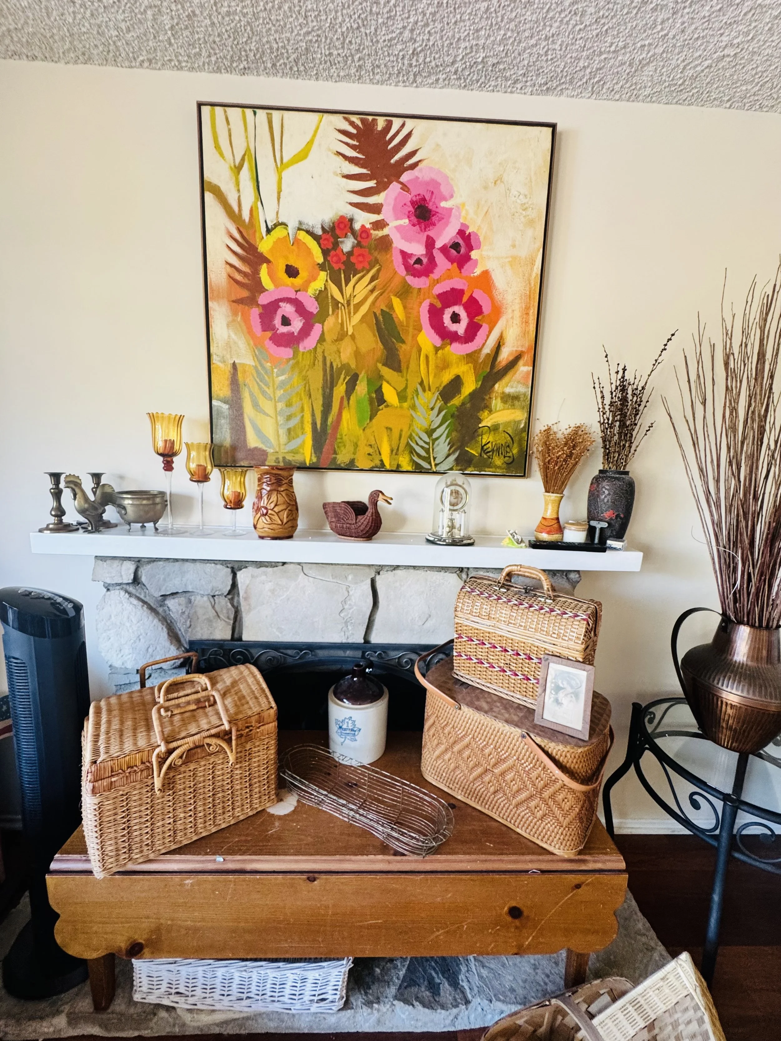 A living room corner with a white mantel and stone fireplace, decorated with various vases and sculptures, a large colorful floral painting on the wall, and baskets on a wooden table in the foreground.