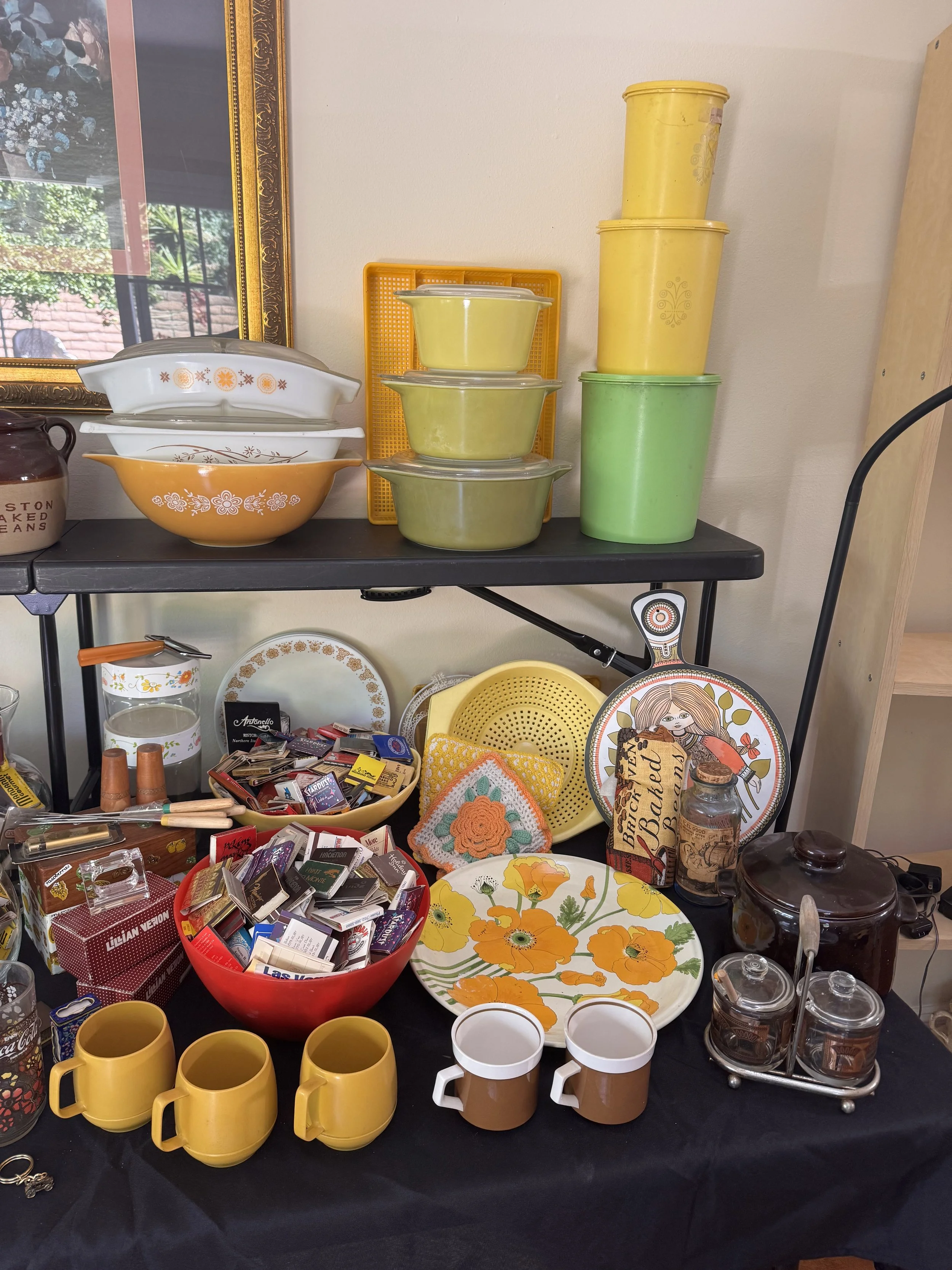A table displaying various kitchenware and household items, including yellow mugs, ceramic bowls, vintage dessert dishes, a floral plate, and a collection of small items, with a mirror and boxed items in the background.