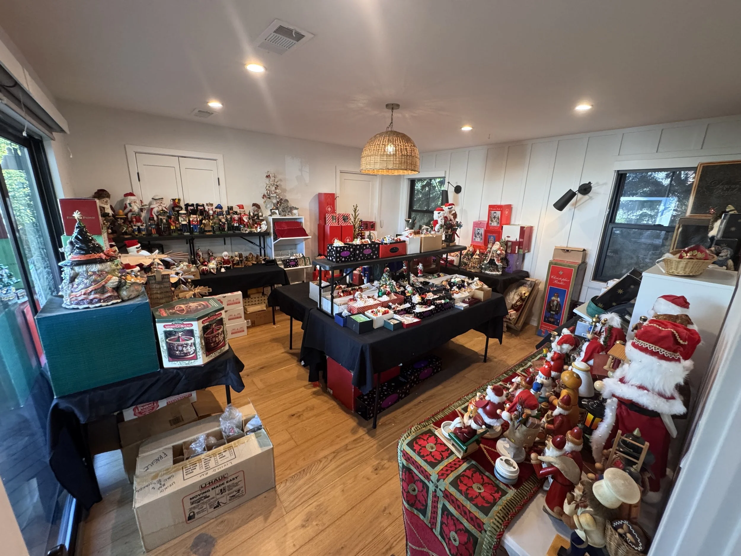 Room decorated with Christmas items including Santa figurines, ornaments, and holiday-themed decorations displayed on tables and shelves, with a window and door in the background.