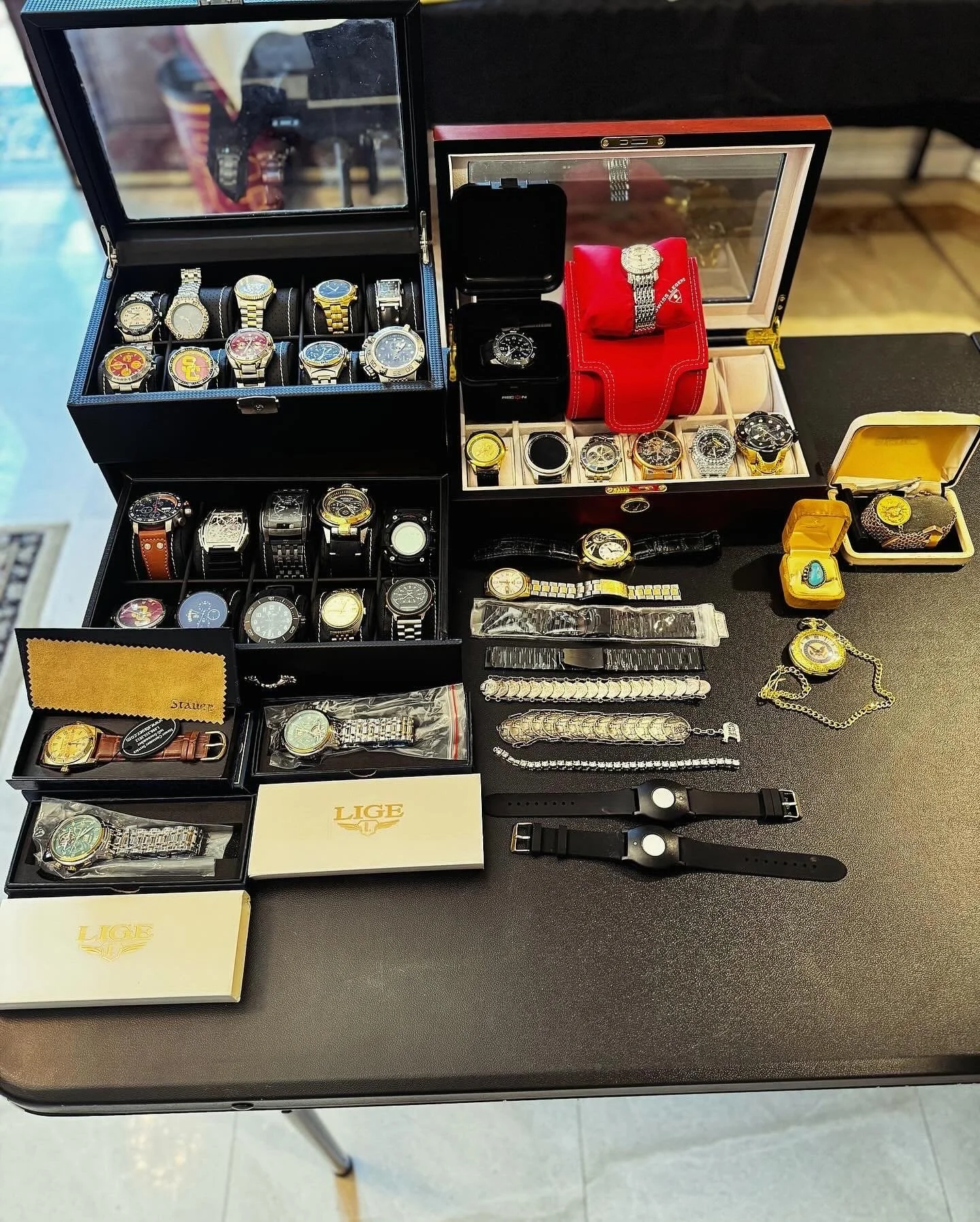Collection of various wristwatches in watch boxes and on display on a table, some with metal bands, leather bands, and digital features, along with watch accessories and jewelry.