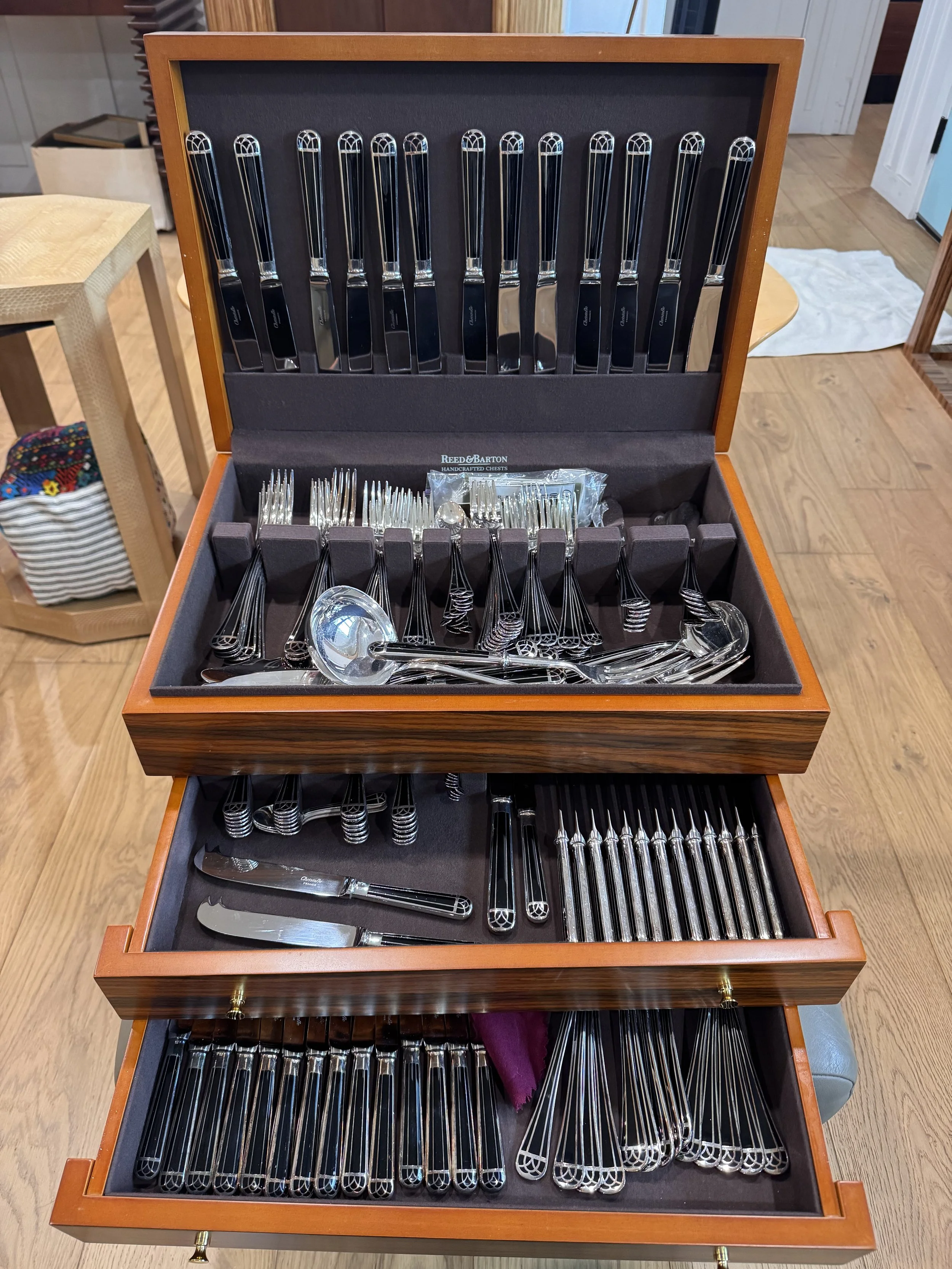 A wooden jewelry box with three drawers, containing a variety of silverware including forks, knives, spoons, and serving utensils organized in separate compartments.