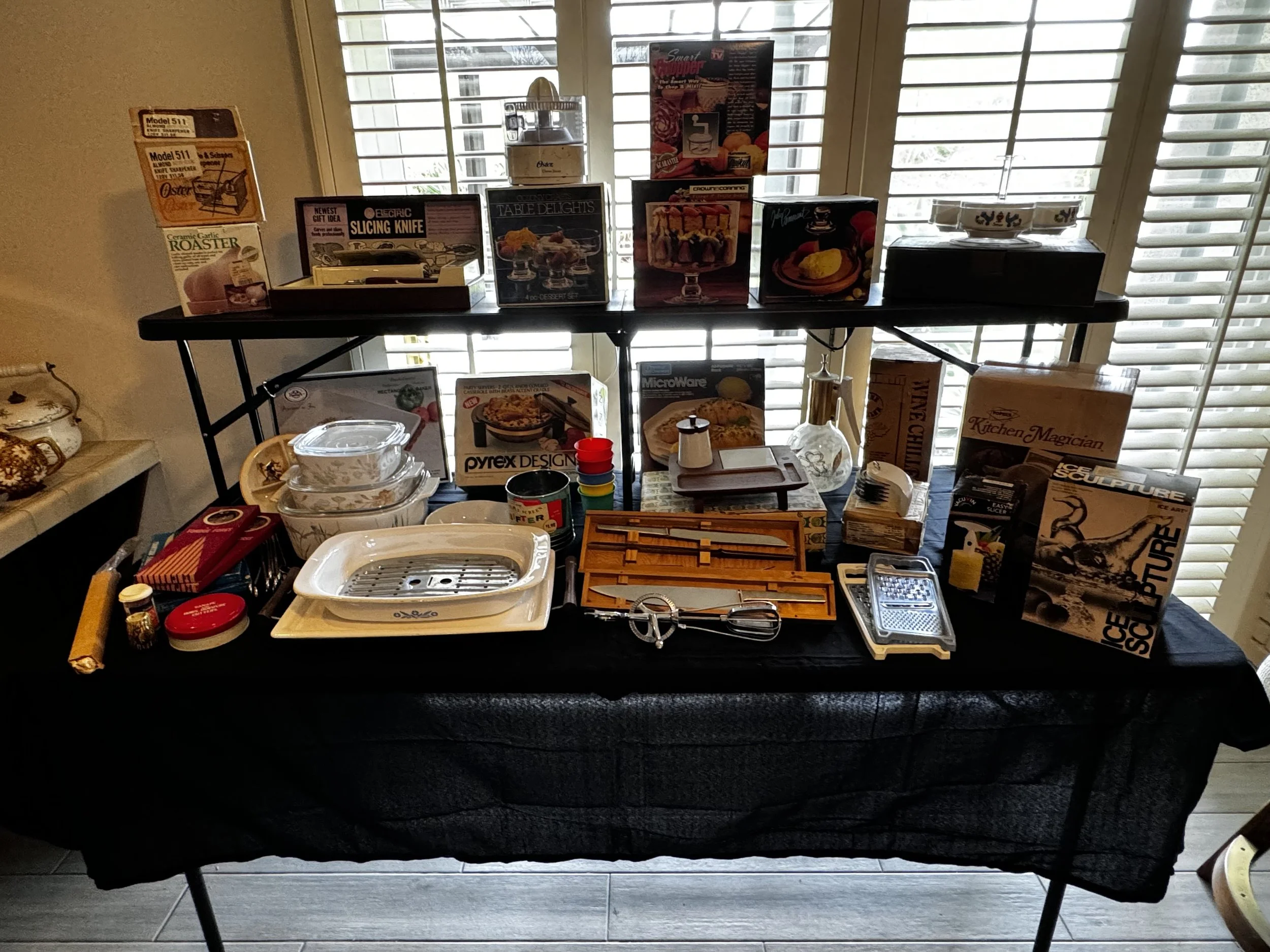 A black table covered with a black tablecloth displaying various kitchen and cooking items, including boxes of kitchen tools, a white ceramic bowl, small containers, kitchen utensils, and cookbooks, set in front of a window with open blinds.