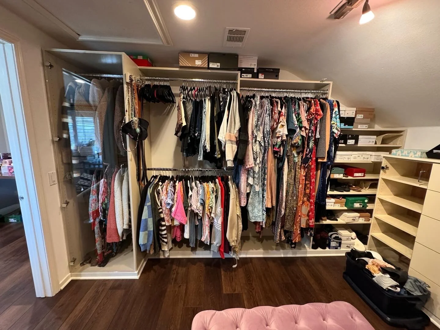 A walk-in closet with clothing on racks and shelves. The clothing includes dresses, shirts, and jackets. There are shoe boxes on top of the shelves and a suitcase on the floor with clothes inside. The room has hardwood floors and a pink cushioned ott