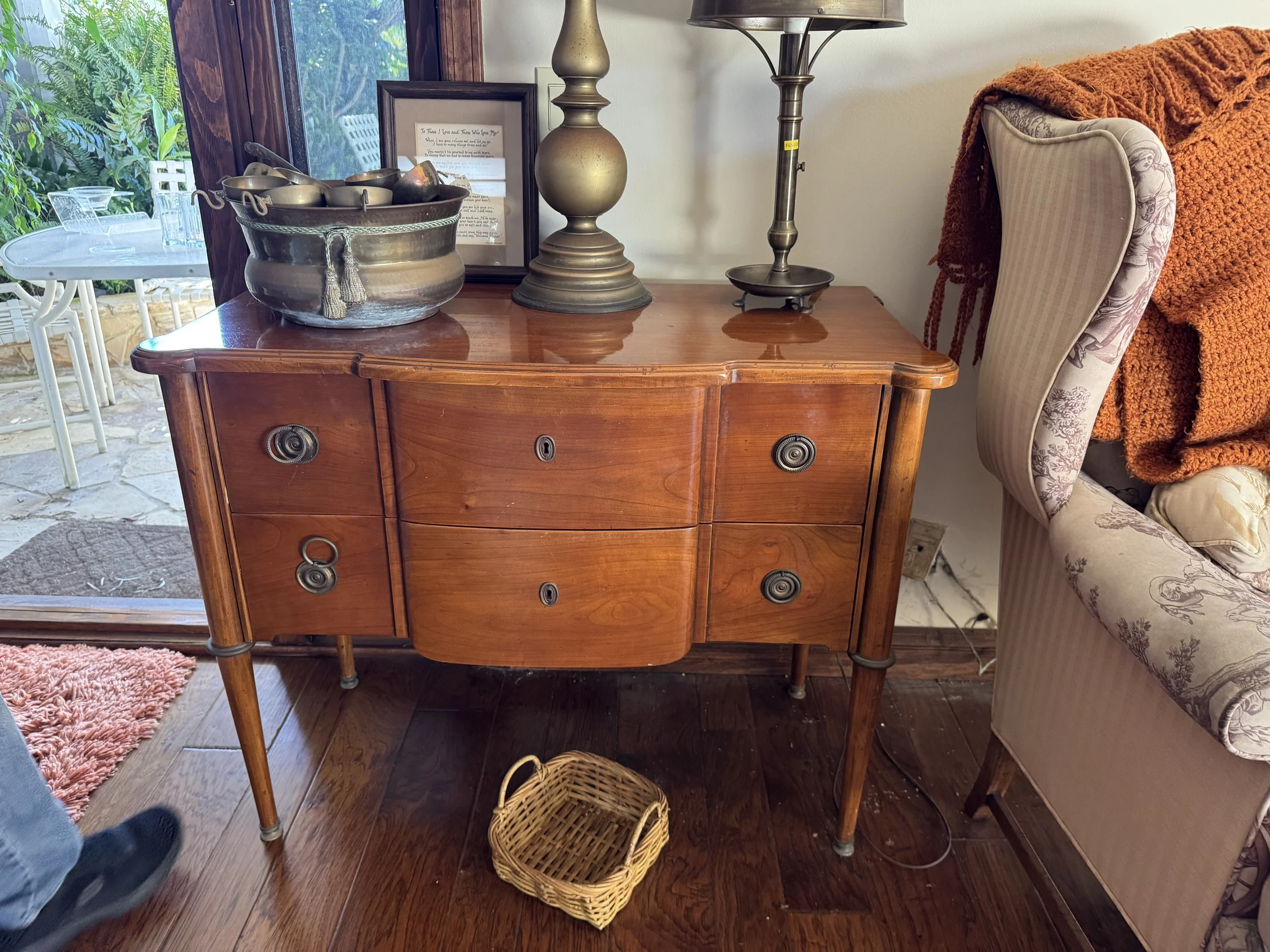 A wooden sideboard with a large metal bowl filled with small metal bowls and utensils, a framed handwritten note, a metallic table lamp, and a vintage-style armchair partially covered with an orange blanket. There is a window behind the sideboard rev