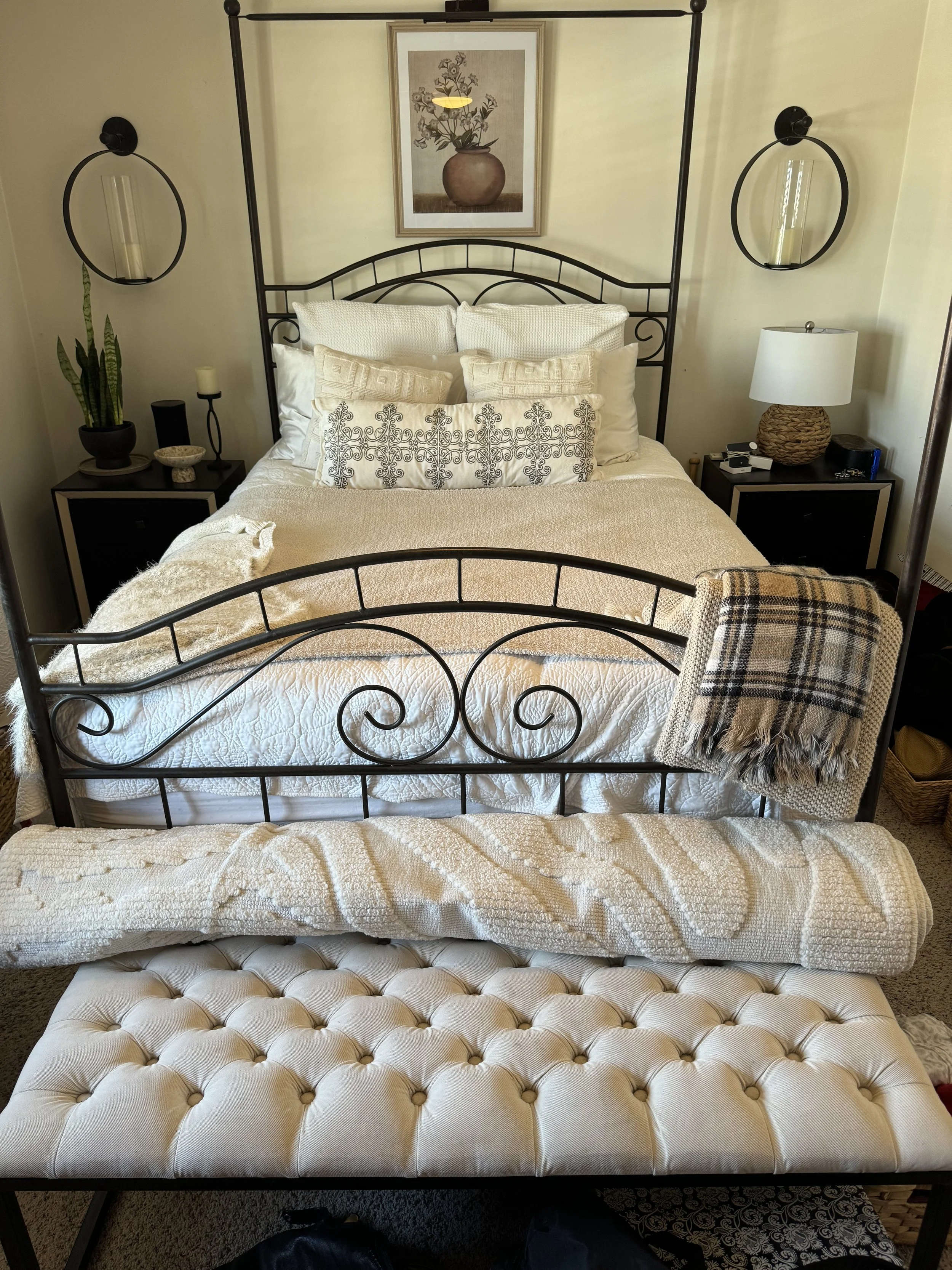 A bedroom with a metal bed frame, white bedding, and multiple pillows. There are two black nightstands with lamps and decor, a framed floral print above the bed, and a cream-colored tufted ottoman at the foot of the bed. A plaid blanket is draped ove