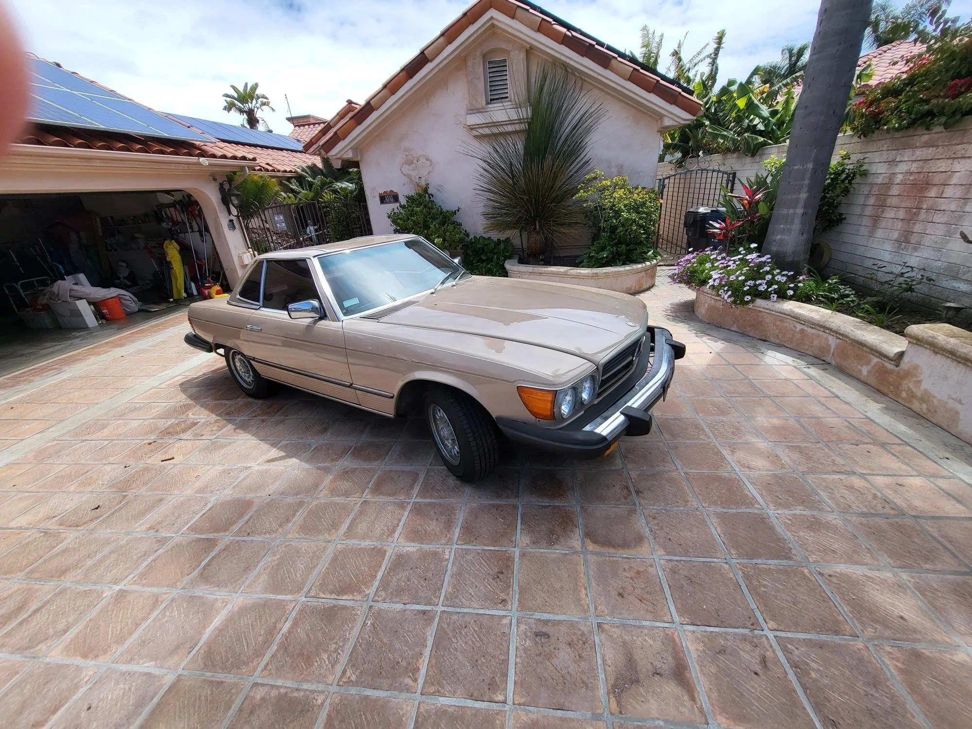 A beige vintage Mercedes-Benz parked on a tiled driveway in front of a white house with a red tile roof, surrounded by lush green plants and colorful flowers.