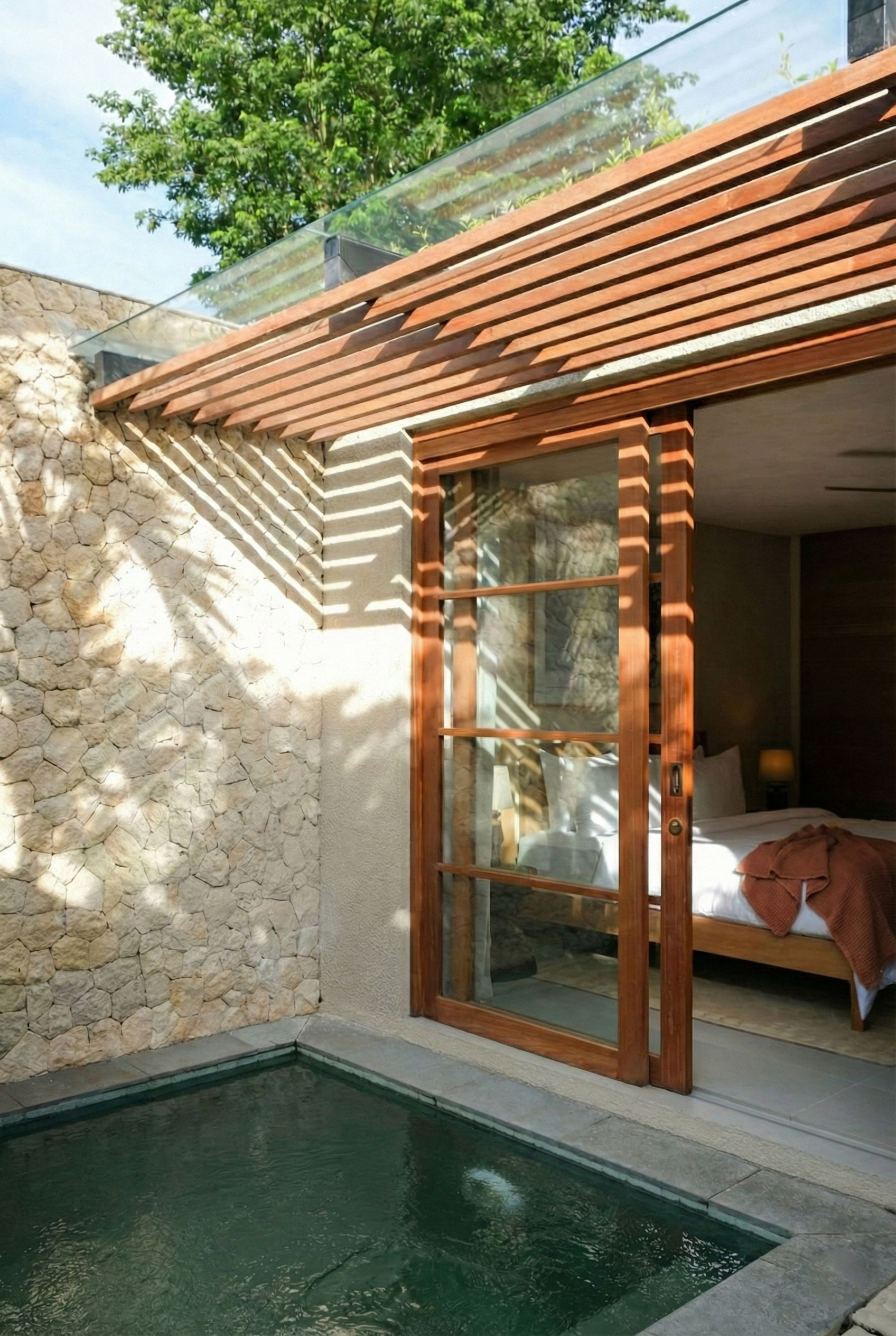 Modern house exterior with stone and plaster wall, wooden and glass awning, and a small pool in the foreground. The house has a large sliding glass door leading to a bedroom inside, with a bed and brown blanket visible.