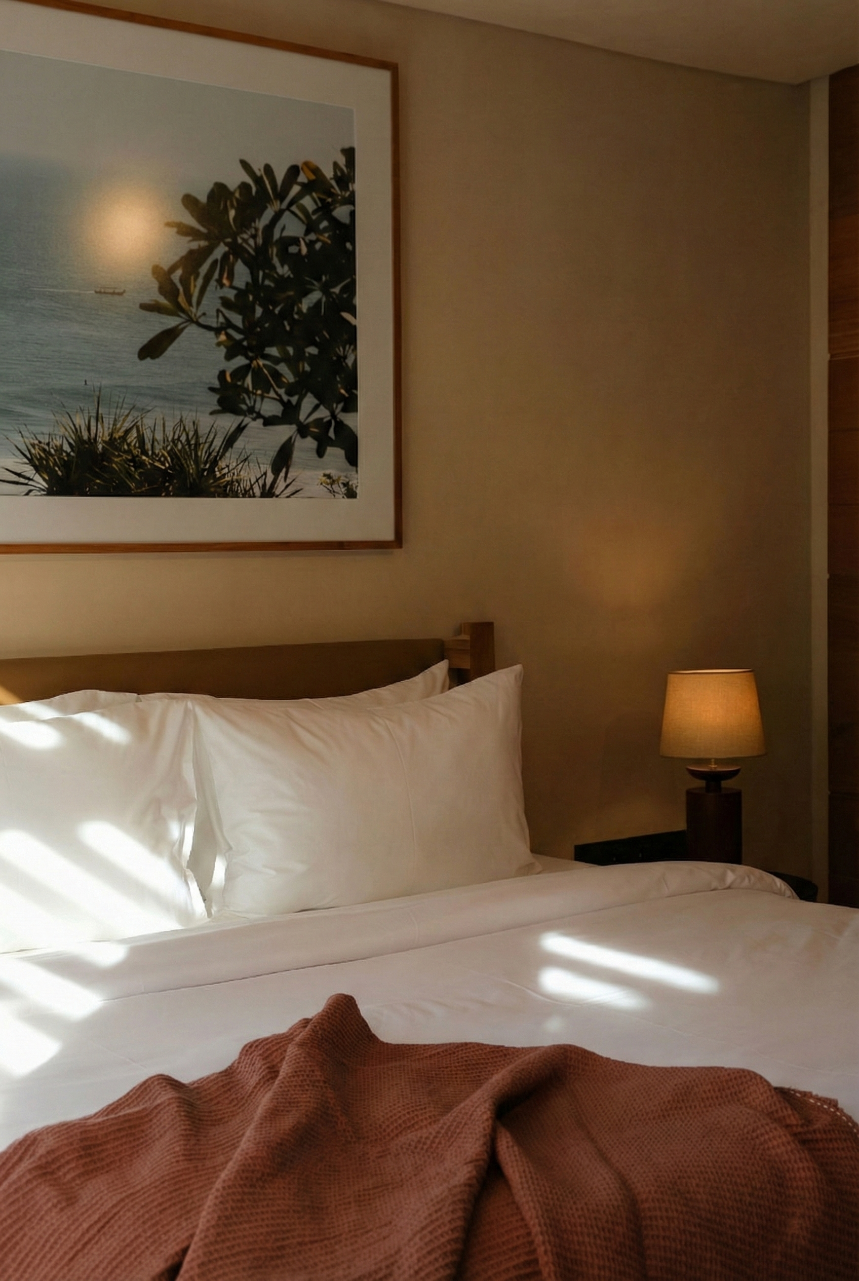 A neatly made bed with white sheets and pillows, a brown throw blanket, and a bedside lamp on a wooden nightstand. A large framed photograph of a tree and ocean scene hangs above the bed on a beige wall.