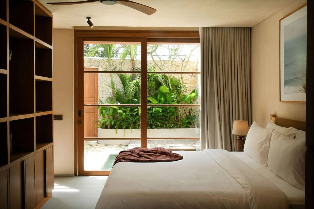 A cozy bedroom with a large bed, white linens, and a bedside lamp, overlooking a lush outdoor garden through a glass sliding door.