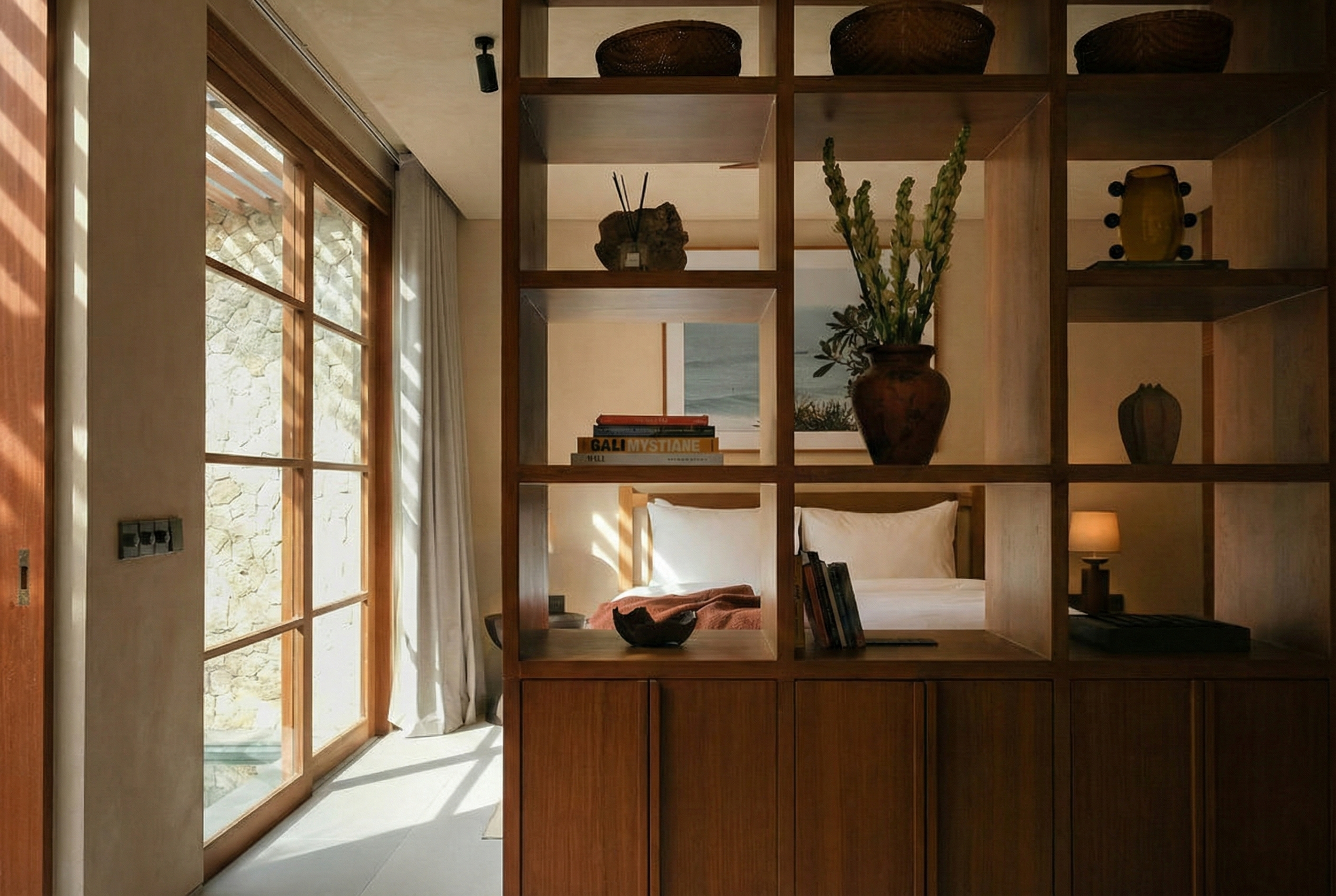 Interior view of a bedroom separated by a wooden room divider with shelves. The bed has white linens and a wooden headboard. There are books, decorative objects, and a large vase with green plants on the shelves. Sunlight comes through large windows 
